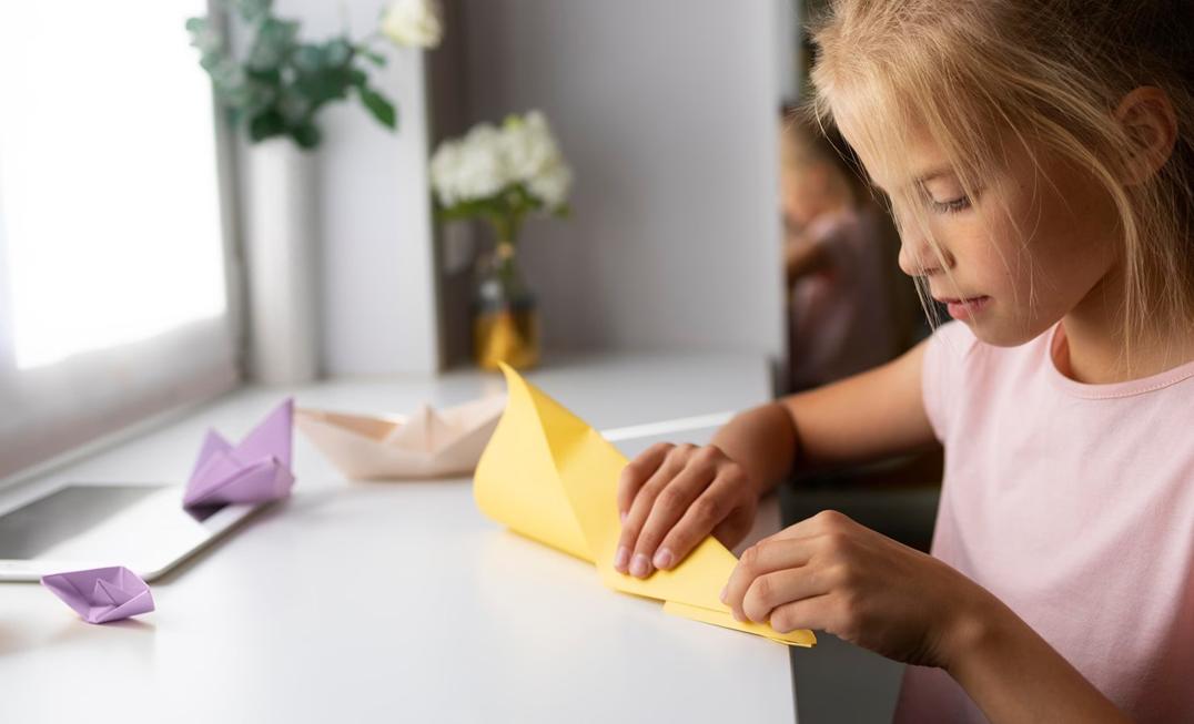 A blonde girl, wearing a pale pink t-shirt, sits at a white table, attentively folding a piece of yellow paper into a boat shape, with other completed paper boats visible nearby.