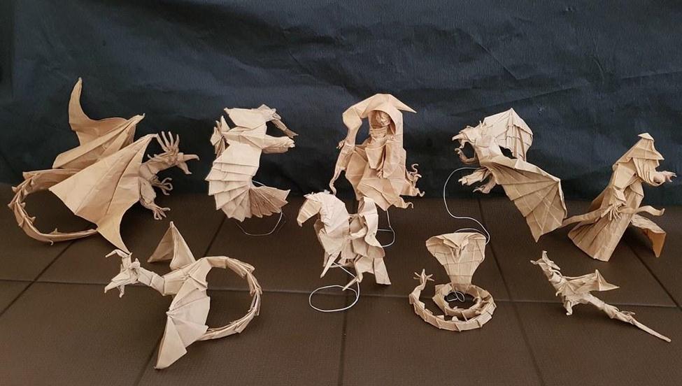 A collection of intricately folded origami figures, predominantly dragons, a hooded figure, a horse, and a cobra, are displayed on a dark surface, showcasing the artist's skill and creativity in paper folding.