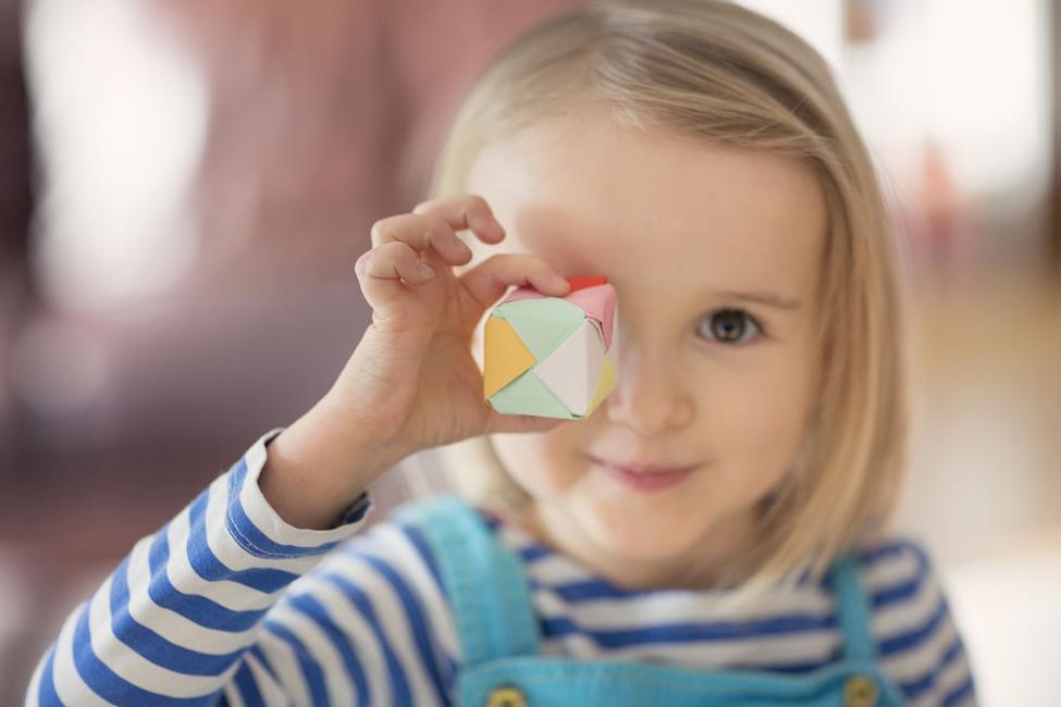 A young girl with blonde hair, wearing a blue and white striped top, playfully holds a small, colourful origami cube up to her eye, a charming and engaging image.