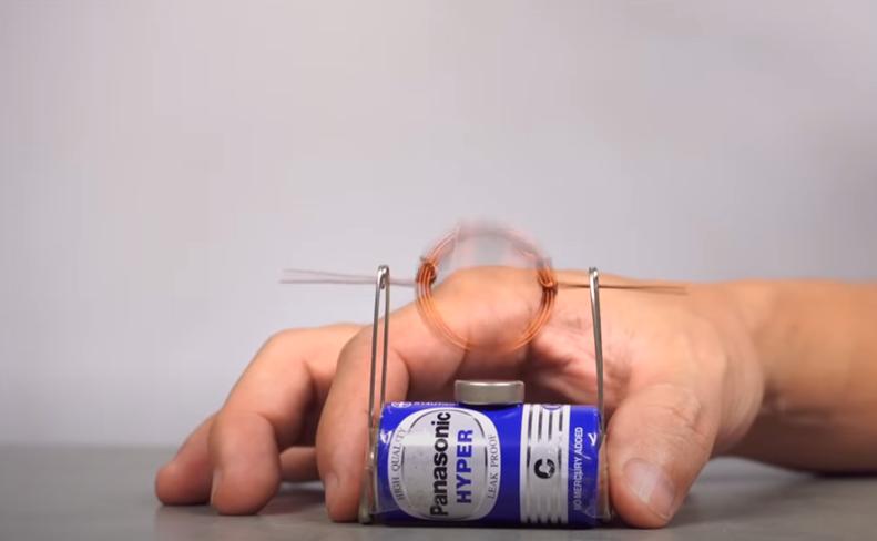 A hand supports a simple homemade electric motor; a coil of copper wire spins rapidly atop a battery and magnet, demonstrating basic electromagnetic principles.