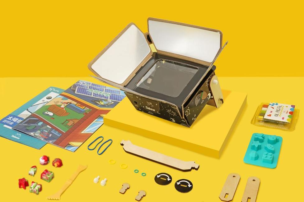 A flatlay image shows a disassembled children's STEM kit on a yellow surface. The main component is a dark cardboard projector with its cover open. Surrounding it are various small parts, coloured pencils, and instruction booklets, suggesting a creative, hands-on construction project.