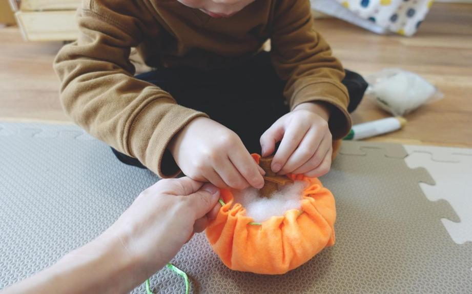 A young child, wearing a brown sweatshirt, carefully inserts a small wooden piece into a partially completed orange felt pumpkin, assisted by an adult hand guiding the process; a collaborative craft project in progress.