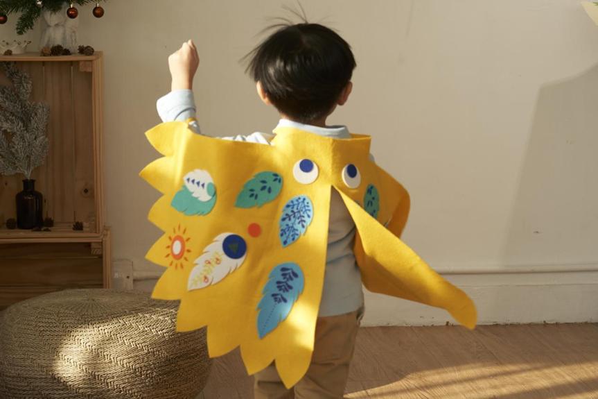 A toddler, seen from behind, proudly wears a handcrafted yellow felt bird costume featuring colourful appliquéd details, suggesting imaginative play and creative self-expression.