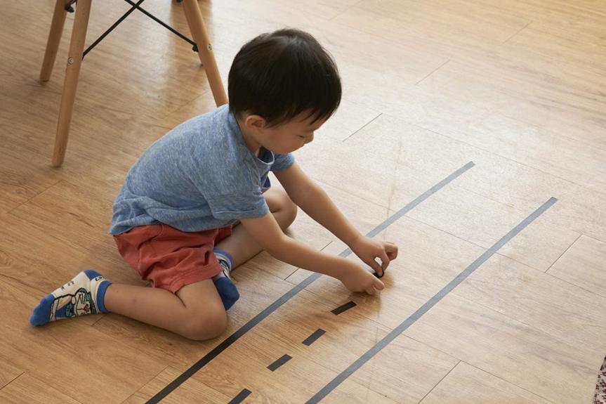 A young boy, wearing a blue t-shirt and red shorts, sits on a wooden floor, playing with a small toy car on a road marked out with black tape; a simple, engaging play scene.