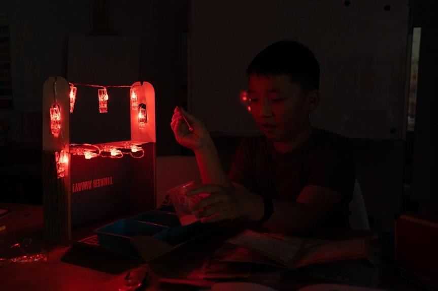 A young person sits in a dimly lit room, illuminated only by a string of red LED lights attached to a small, handcrafted structure; they appear to be working on a project, perhaps a circuit or other creative construction.