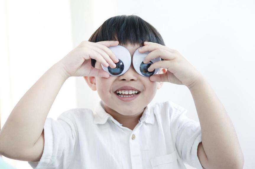 A young boy, wearing a white shirt, holds two large googly eyes over his own, smiling playfully.