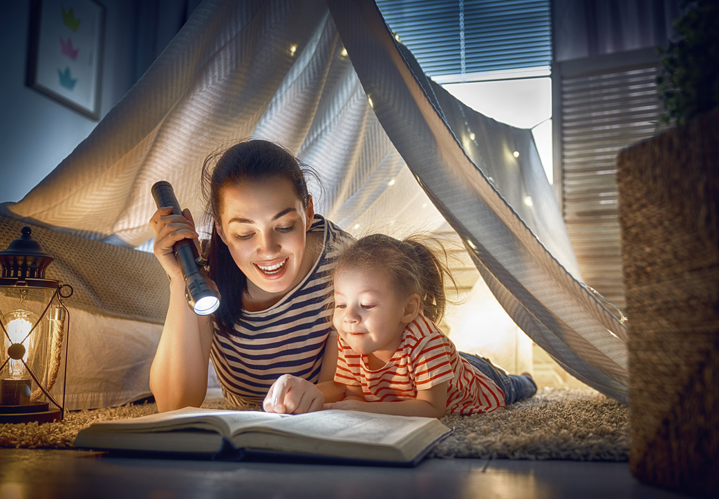 A mother and daughter are snuggled inside a cozy blanket fort, reading a book by the light of a flashlight. The scene is warm and inviting, capturing the joy of shared moments and the magic of Roald Dahl STEM learning.