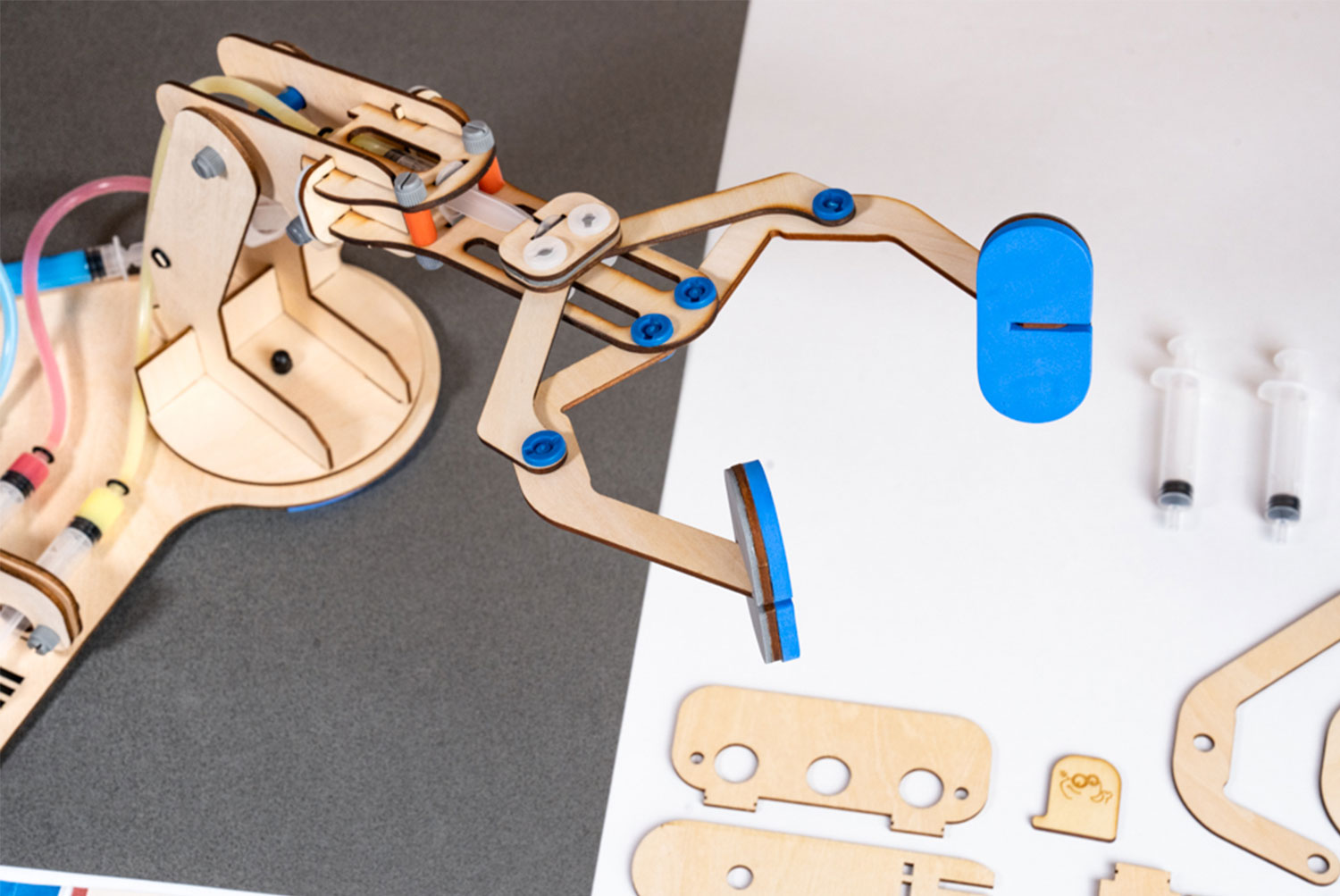 A partially assembled wooden robotic arm, featuring blue padded pincers and clear plastic tubing connected to syringes, is displayed against a split background of dark gray and white. The arm extends over the white section, showcasing its pincers and mechanical components. On the white surface, unassembled wooden parts and two syringes lie near the arm, suggesting a construction kit or a project in progress. The image highlights the educational and hands-on nature of building and experimenting with robotics.