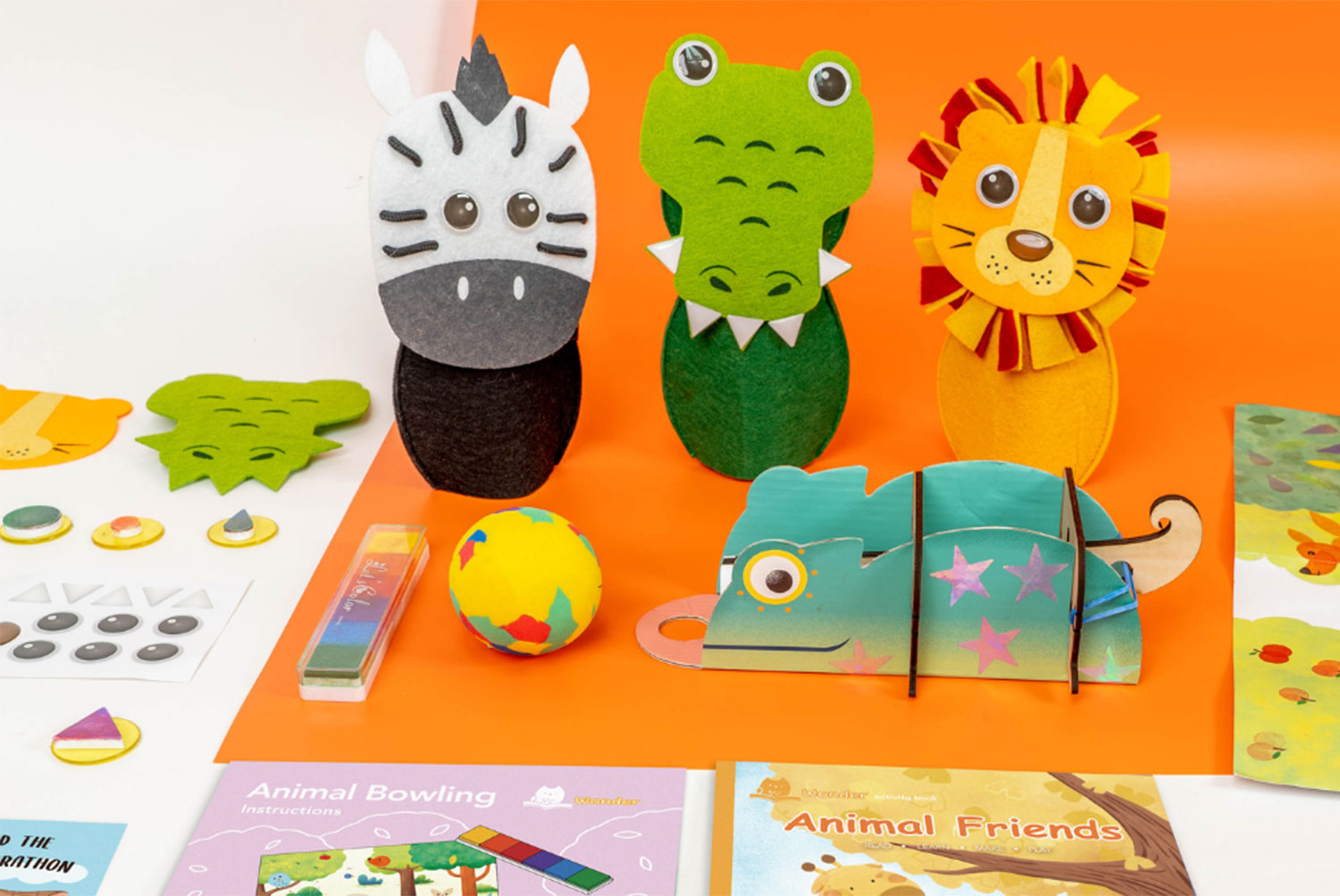 A collection of children's craft and activity items is arranged on a vibrant orange surface, with a portion of a white surface visible in the top left corner. Three felt animal bowling pins, representing a zebra, a crocodile, and a lion, stand upright. A partially assembled cardboard chameleon, a multicolored ball, stickers, felt cutouts, an ink pad with a rainbow gradient, and two small booklets titled Animal Bowling Instructions and Animal Friends are also scattered across the surface. The scene suggests a playful and creative activity kit for children with an animal theme.