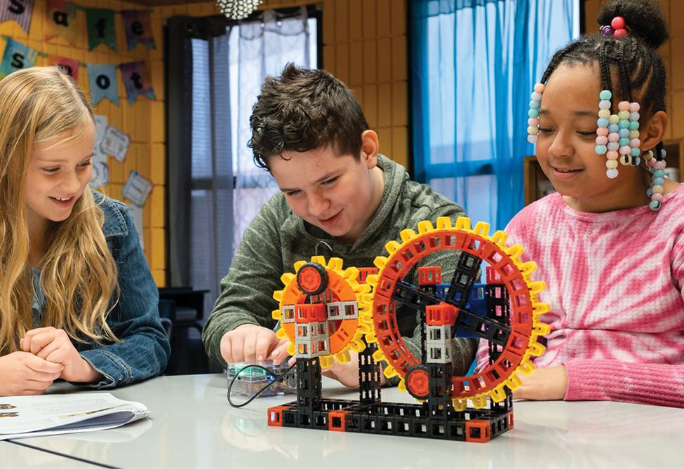 Three children, two girls and a boy, work together on a construction project involving colourful gears and blocks in a classroom setting.