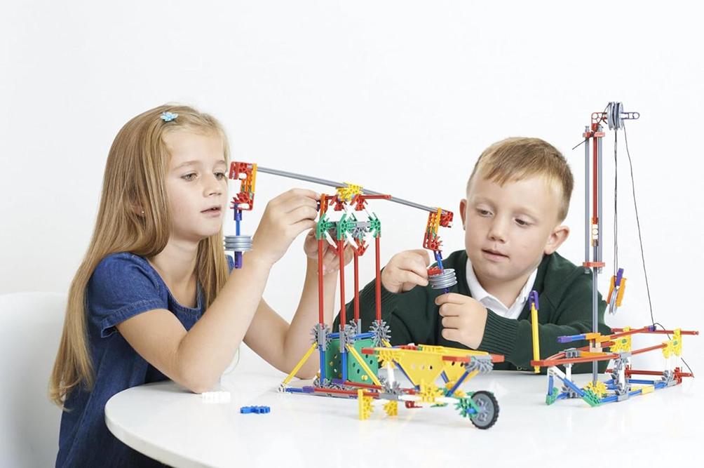 Two young children, a girl and a boy, are engrossed in building a colourful and complex structure using a construction toy set at a white table.