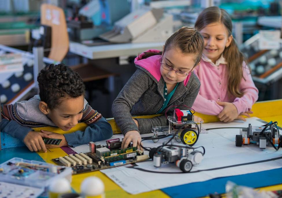 Three young children, two girls and a boy, are engrossed in a robotics project, working with small robots and electronic components on a table in a classroom or workshop.