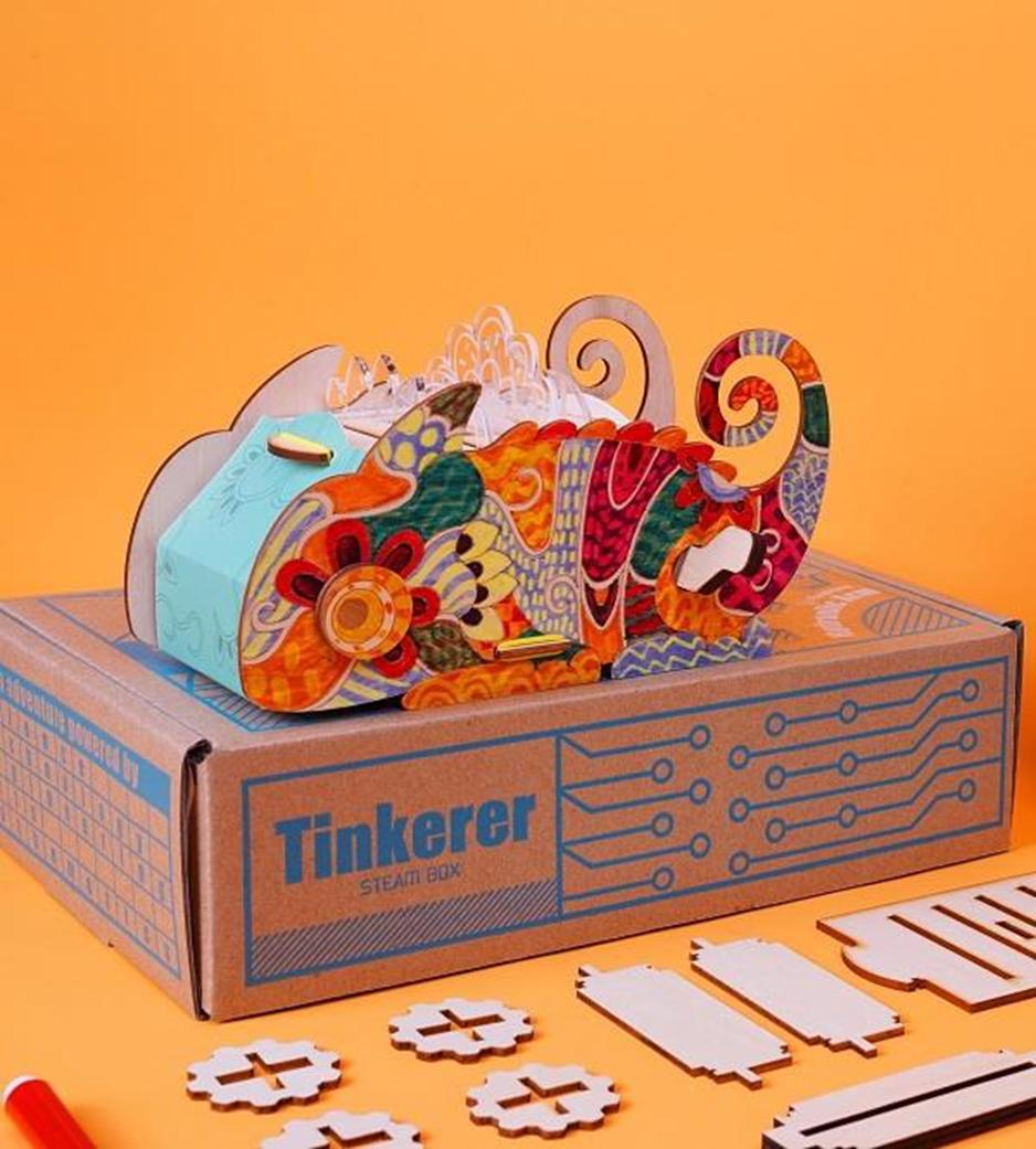 A brightly coloured, patterned chameleon, crafted from wood and possibly other materials, sits atop its box, labelled Tinkerer Steam Box, surrounded by unassembled wooden components.