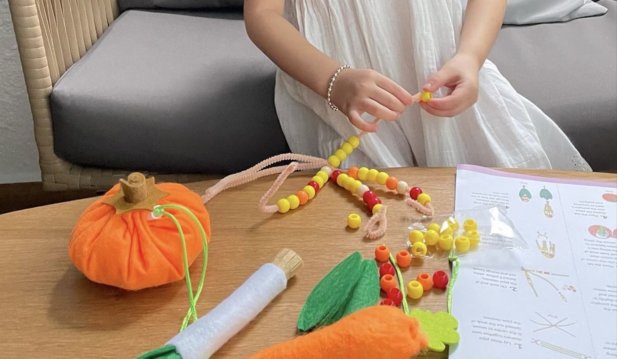 The image depicts a child's hands engaged in a hands-on, creative STEM activity involving various colourful, textured elements. The person appears to be manipulating a collection of beads, shapes, and other tactile materials on a wooden surface, suggesting an artistic or educational STEM endeavor. The arrangement includes an orange fabric pumpkin-shaped object, as well as other geometric shapes and patterns in vibrant hues of yellow, orange, and green. The child's focused attention and the array of materials indicate this is likely a crafting or learning activity, perhaps involving fine motor skills, color recognition, or creative expression. The overall scene captures a moment of engaged, hands-on exploration and creation, with the child's actions and the materials on the table suggesting an immersive, sensory-driven experience. 