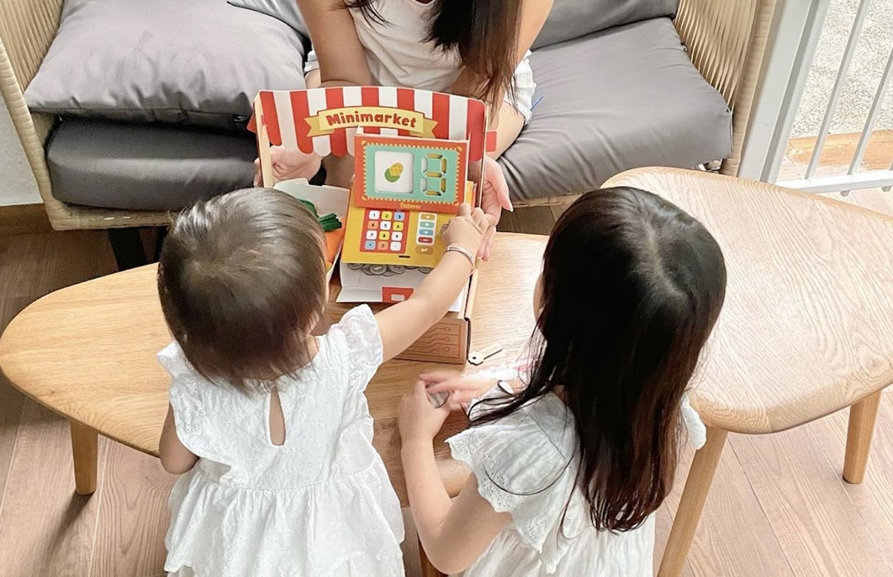 The image shows two children engaged in imaginative play with a miniature market set which is part of the Mini-Mart STEM box. The play set includes a small storefront, shelves stocked with colorful play food and household items, and a cash register.