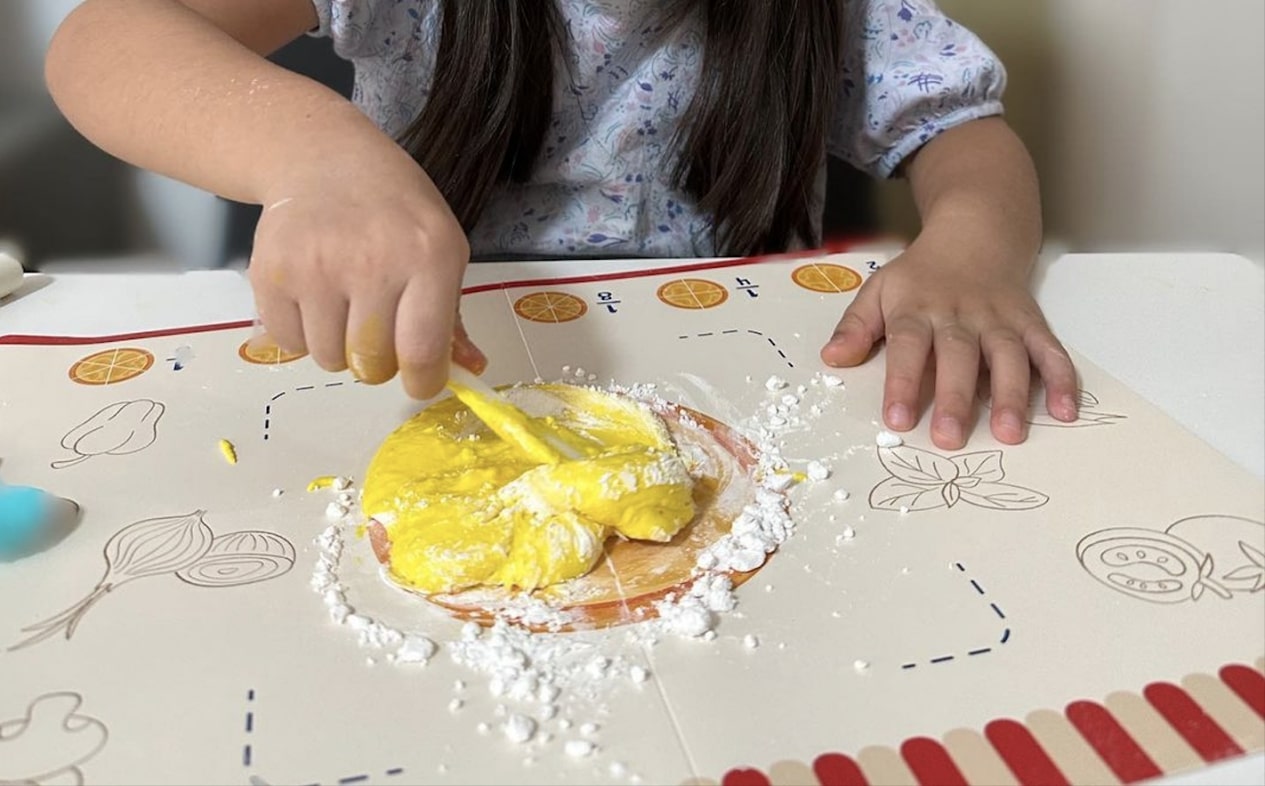 The photo shows a child's hands working with some play dough from the Pizza Maths STEM box, which has a vibrant yellow color. The child is cutting the dough with a plastic knife. The image captures a moment of creative STEAM exploration and sensory engagement.