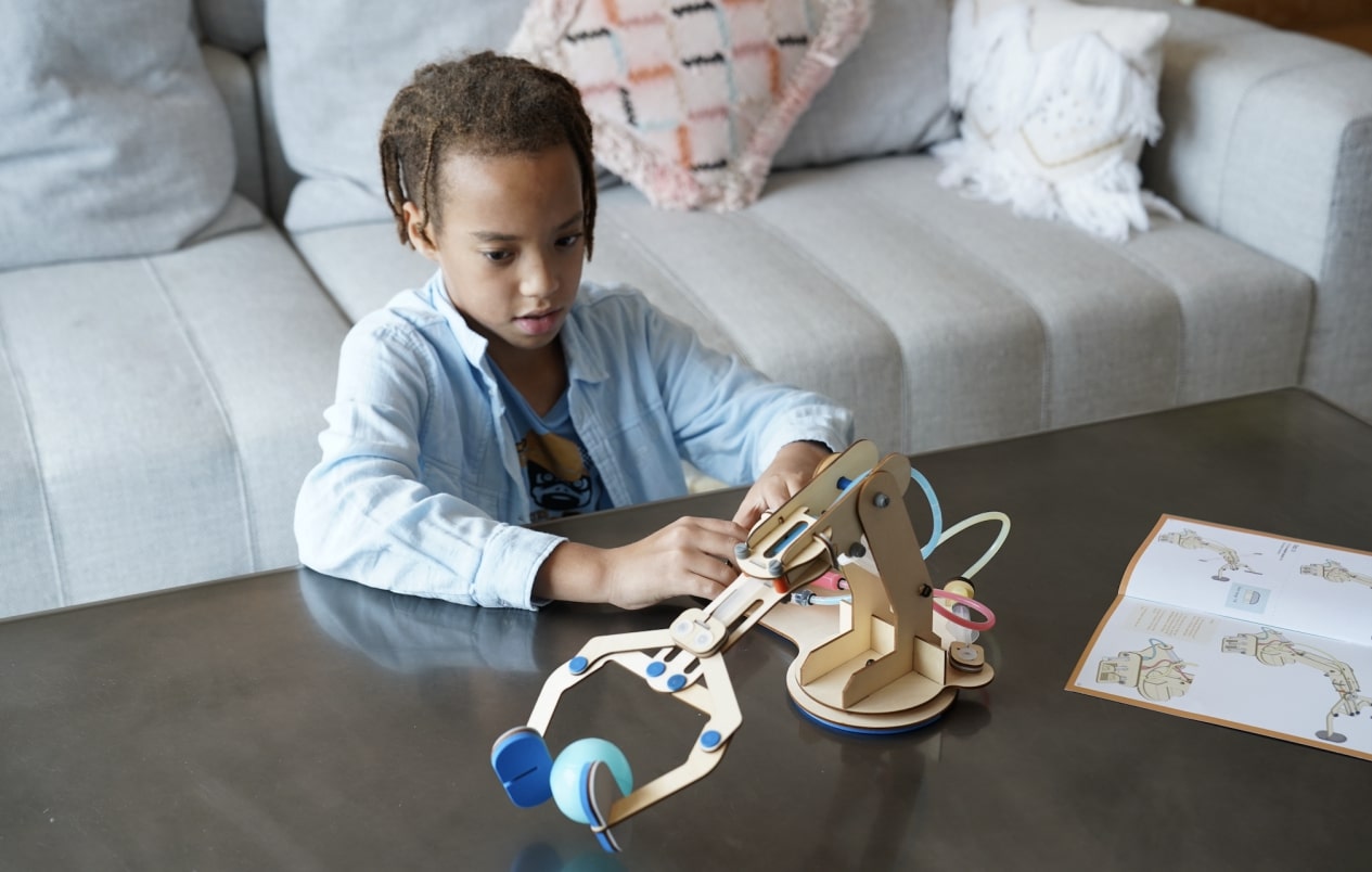 The photo shows a child, intently focused on manipulating a hydraulic arm. The child's expression conveys a sense of concentration and curiosity as they explore the various components and mechanisms of the toy. The setting appears to be a comfortable living room environment, with soft furnishings and decorative elements in the background. The child's engagement with the toy suggests a playful, educational STEM activity that allows them to develop their problem-solving and fine motor skills. The overall scene captures a moment of thoughtful, hands-on learning and exploration.