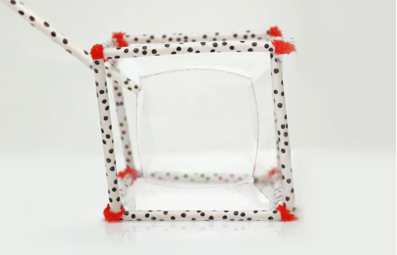 A large, shimmering soap bubble is partially enclosed within a cube constructed from white paper straws with black polka dots. The corners of the cube are connected with bits of red pipe cleaner. A single straw with the same polka dot pattern enters the frame from the upper left, seemingly interacting with the bubble. The entire structure rests on a white reflective surface.