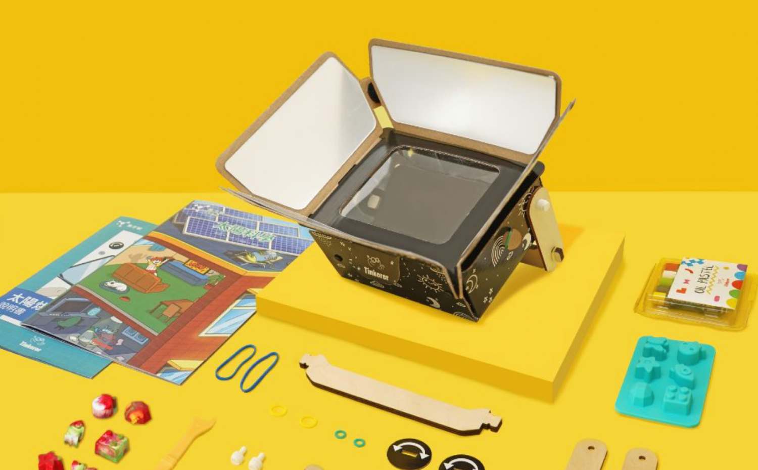 A Tinkerer branded DIY projector kit is displayed open, showcasing its cardboard structure with a black front panel featuring a clear window and space-themed doodles. The projector's hinged top flaps are open, revealing a reflective silver interior. Surrounding the projector on the bright yellow surface are various kit components, including illustrated instruction booklets, wooden structural pieces, rubber bands, small rings, oil pastels, a teal silicone mold with shapes, and small, colorful decorative items. The arrangement suggests the kit is ready for assembly.