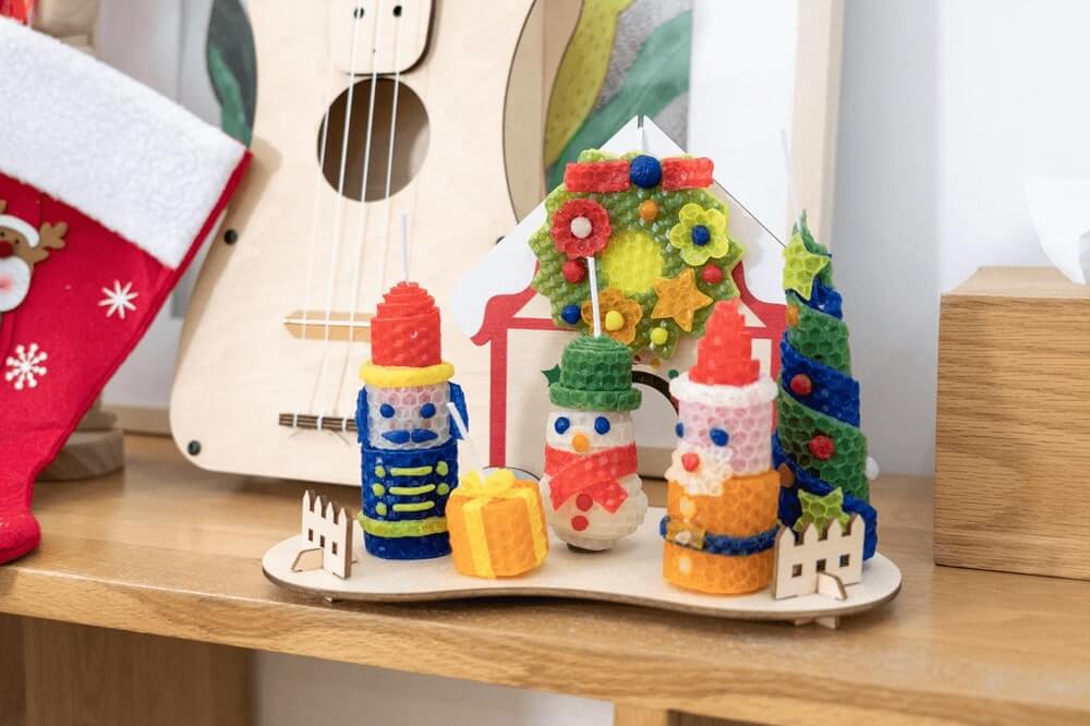 Several small decorative Christmas figures made from textured, colourful wax sit on a wooden display stand on a shelf, alongside a Christmas stocking and a ukulele. The wax figures include a nutcracker, a snowman, a Santa Claus, a Christmas tree, and a Christmas wreath. A small wooden fence and a present-shaped wax figure are also part of the scene.