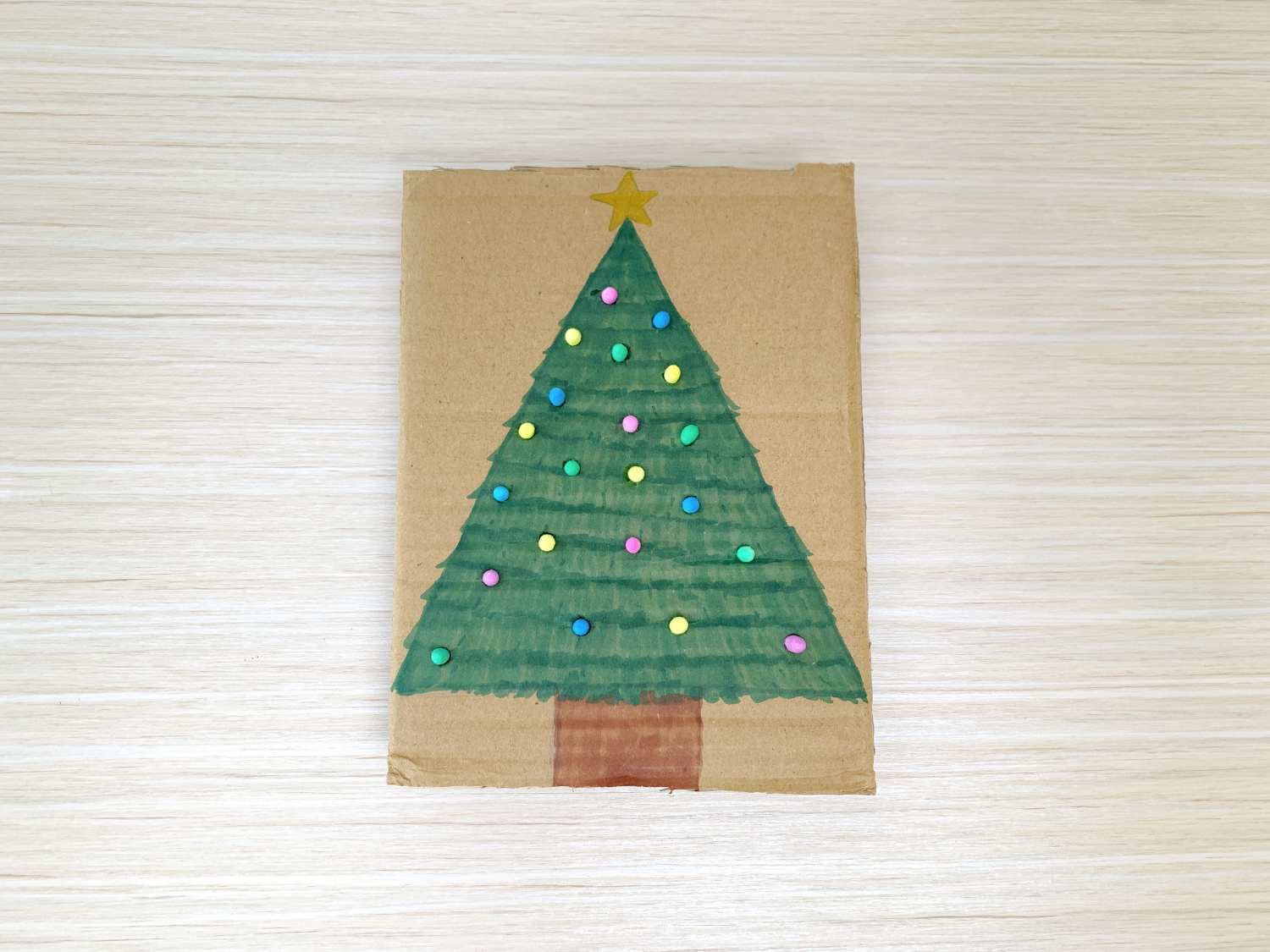 A Christmas tree is drawn and decorated on a piece of cardboard, placed on a light wooden surface.