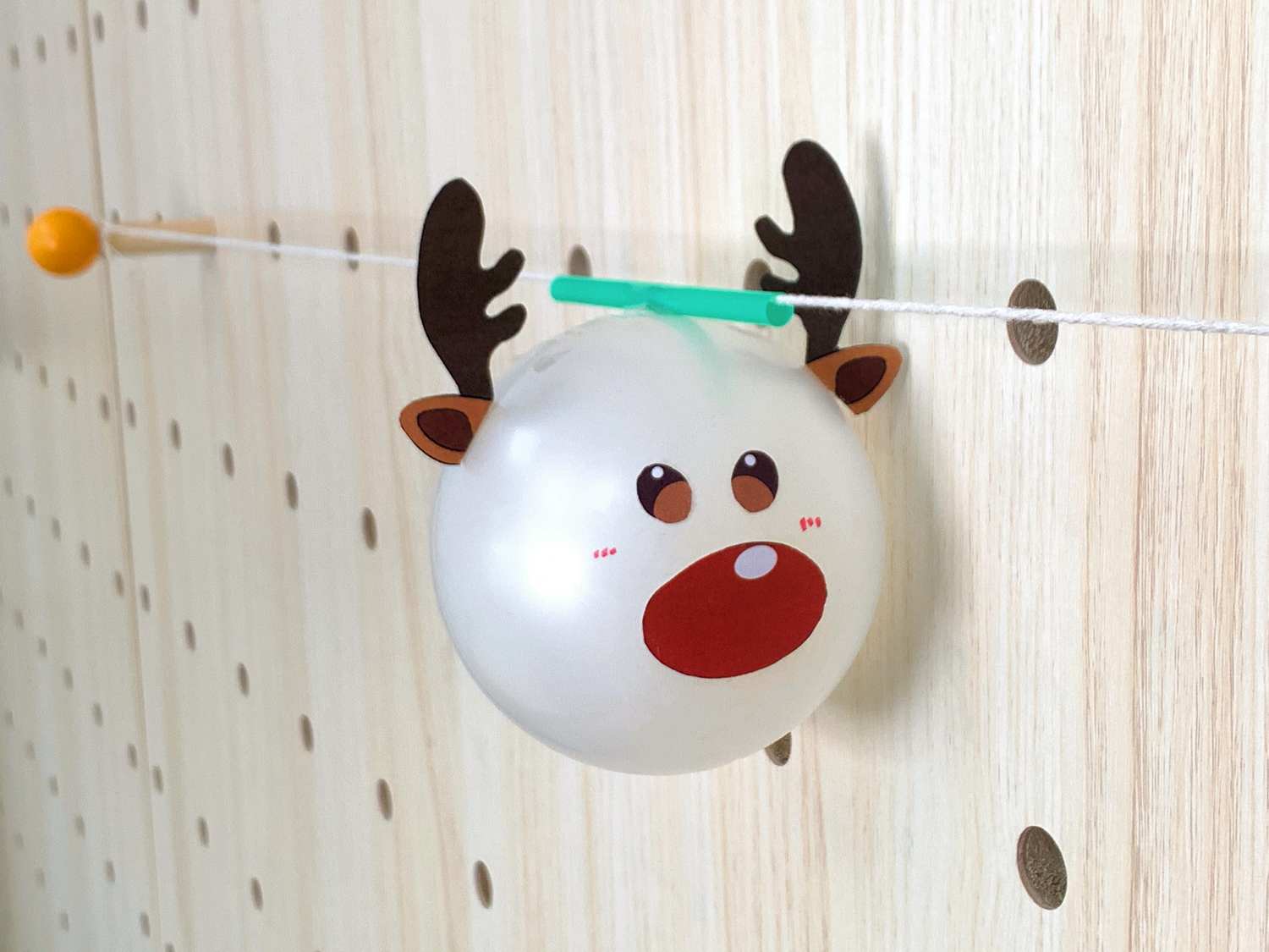 A white balloon decorated with a reindeer face hangs from a string against a pegboard wall.