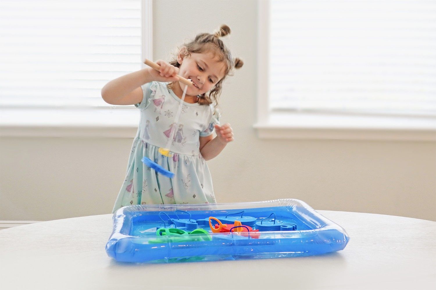 A cheerful young girl in a blue and white dress, her hair styled in two cute buns, engages in a delightful fishing game. She grasps a wooden fishing rod, its line dangling into a blue inflatable tub filled with water. Her face lights up with joy as she focuses on a blue plastic fish hooked to her line, floating in the makeshift pond. The scene captures the essence of childhood play, showcasing the simple pleasures and challenges of a toy fishing set. The girl's beaming smile and concentrated gaze reflect her immersion in this playful activity, embodying the pure, unfiltered happiness that comes from imaginative play and small accomplishments.