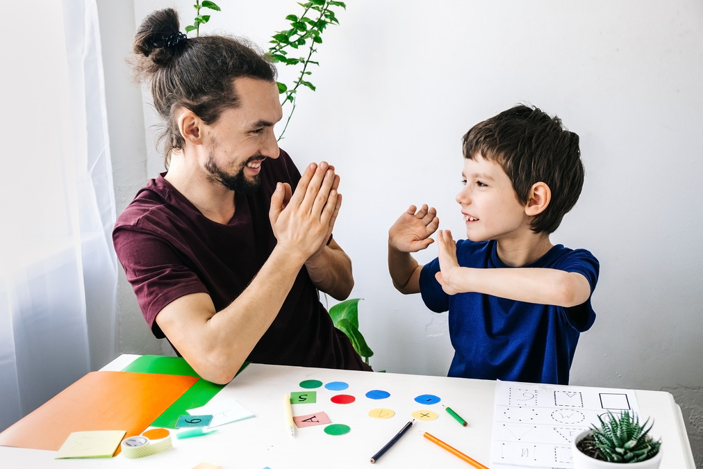 A father and son sit at a table strewn with vibrant sheets of paper and an array of pencils, hinting at an engaging STEM project underway. Their faces are lit up with joy as they take a playful break from their educational pursuit. The father's hands are joined together, while his son's are extended forward, mimicking the classic pose of a patty-cake game. Their shared laughter fills the air, showcasing the beautiful bond between parent and child as they blend learning with fun.