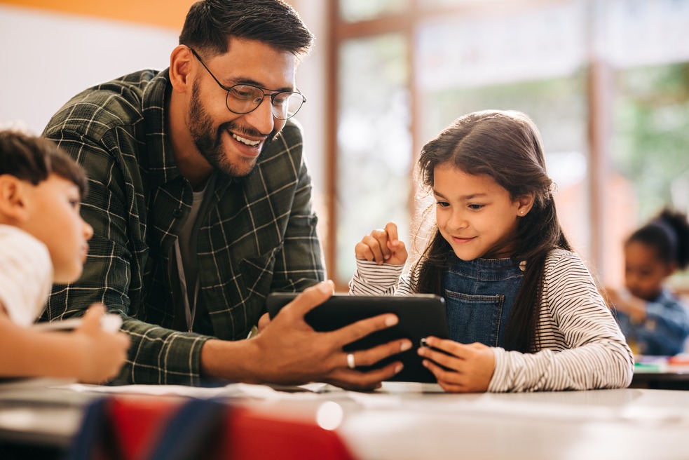 A young girl is back to school in a striped shirt and overalls leans forward, her eyes glued to the screen of a tablet. A smiling teacher, in a plaid shirt, looks on with amusement. The scene is a snapshot of learning in the digital age, with a focus on the teacher's warm and supportive presence.