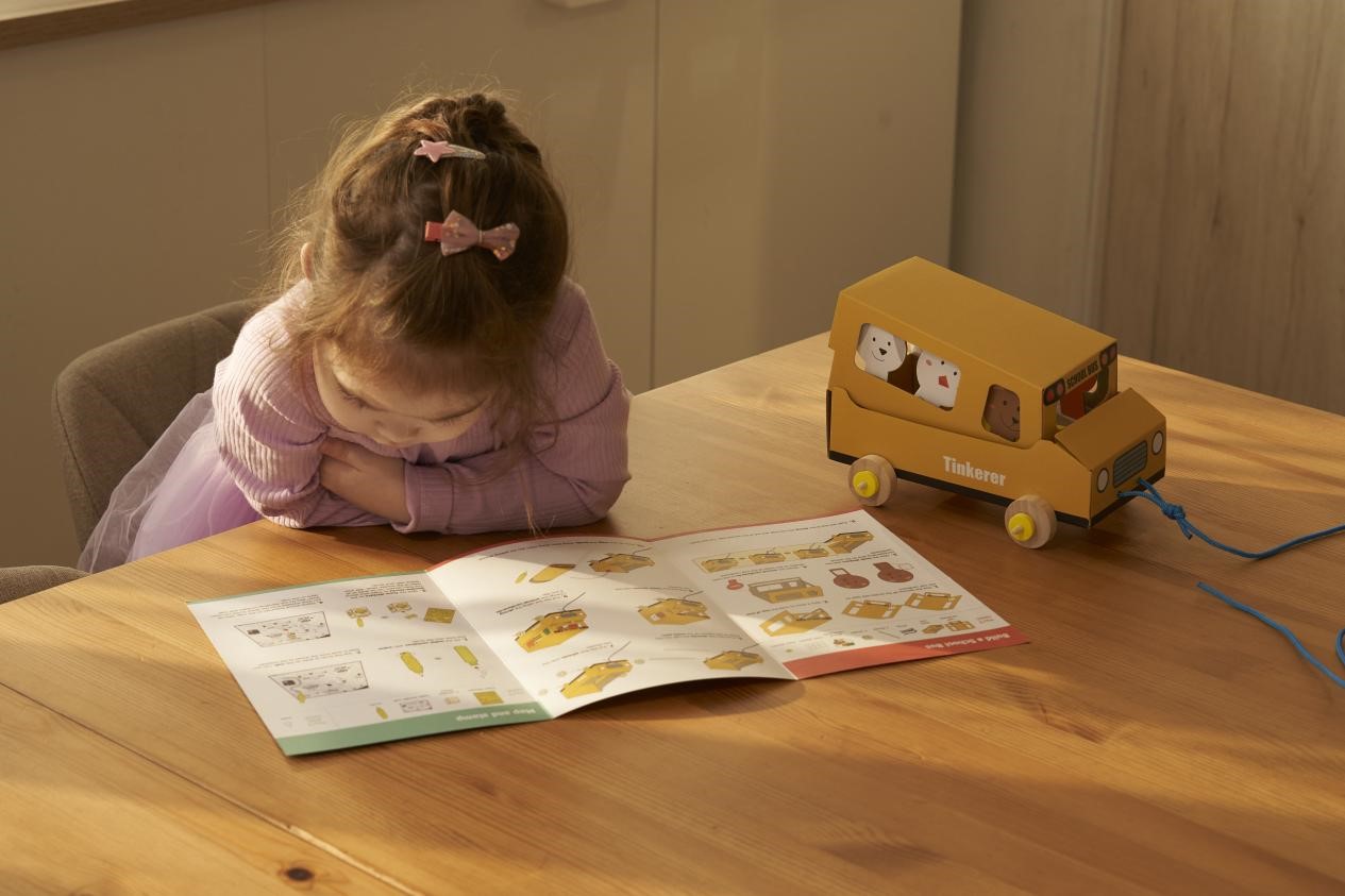 A young girl, with her hair adorned with colourful bows, leans over a table, intently studying the instructions for assembling a toy bus. The bus, painted a cheerful yellow, sits on the table beside her, a testament to the joy of hands-on learning and the magic of creating something new.