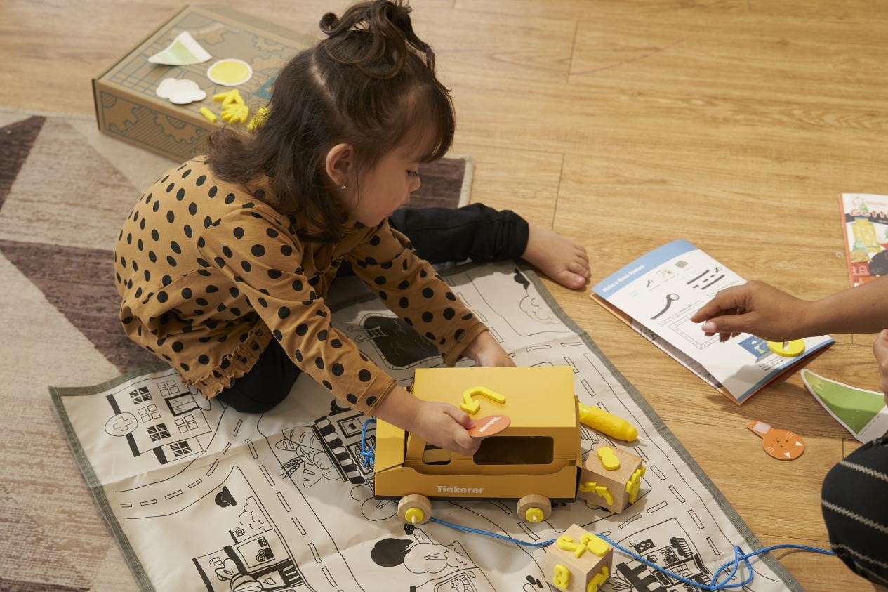 A child in a brown and black polka dot shirt sits on a playmat, engrossed in assembling a yellow wooden toy bus. She holds a yellow piece in her hand, ready to attach it to the bus. The scene is warm and inviting, showcasing the joy of learning and playing through hands-on activities.