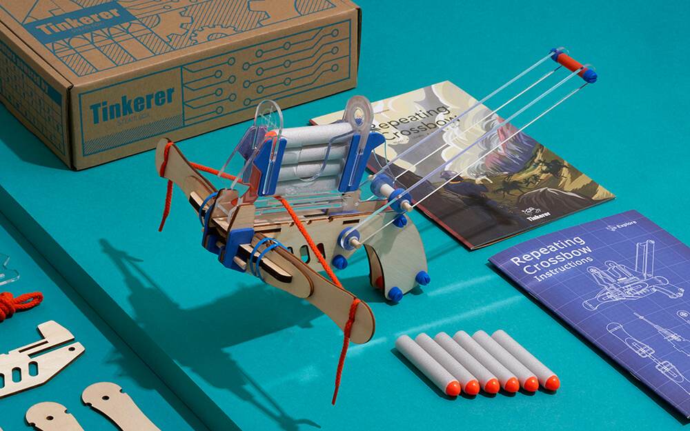An assembled wooden and acrylic toy repeating crossbow from a Tinkerer STEAM kit is displayed on a vibrant teal surface, along with its branded box, instruction manuals, and a row of foam darts with orange tips.