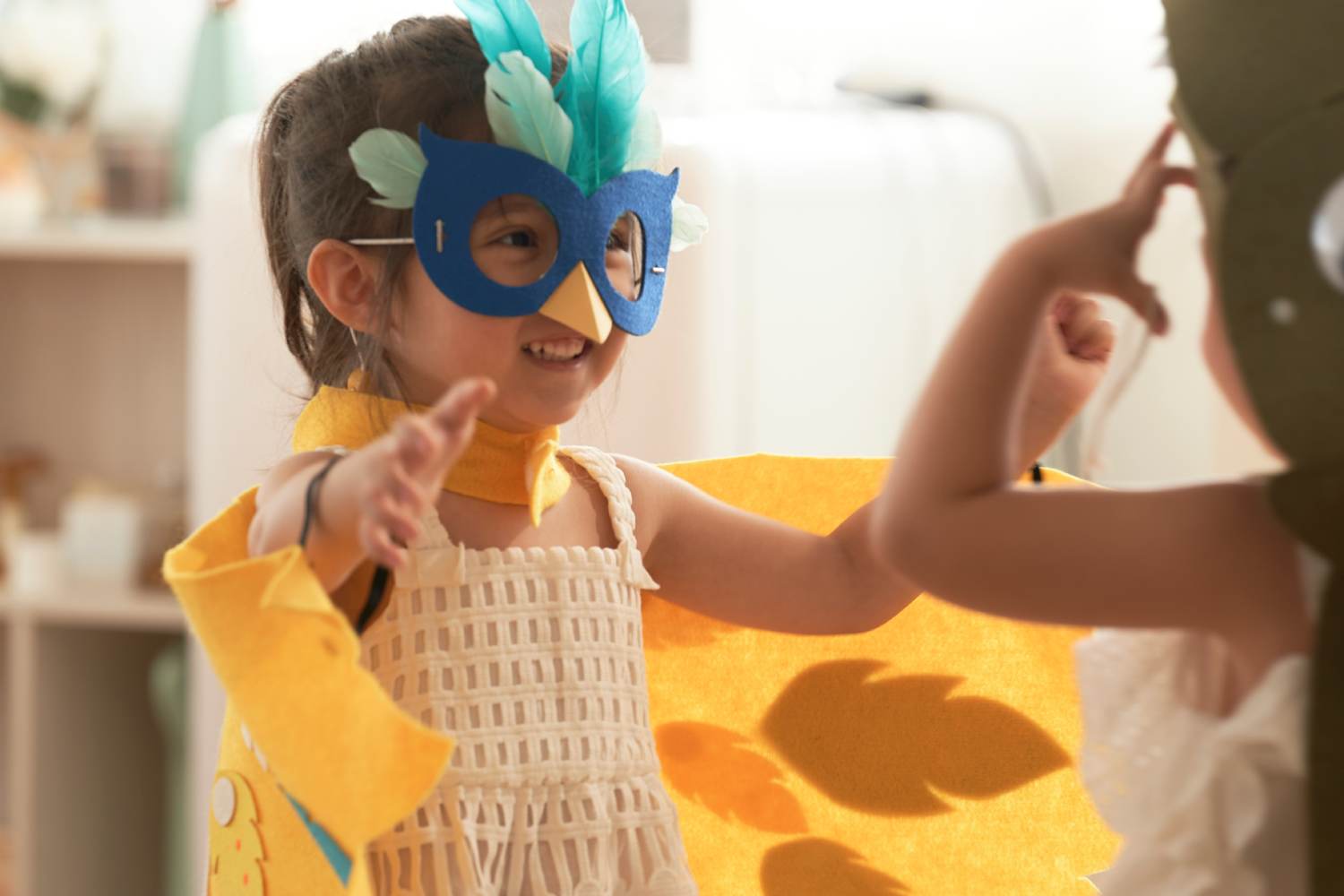 A smiling young girl in a playful, homemade bird costume, complete with a blue feathered mask and a yellow felt cape, interacts with another child who is partially visible on the right.