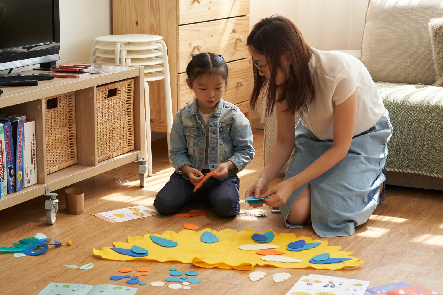A woman and a young girl are kneeling on a wooden floor in a brightly lit living room, working together on a colorful felt craft project. They are arranging blue, orange, and white felt feathers onto a large yellow felt base that is spread out between them.