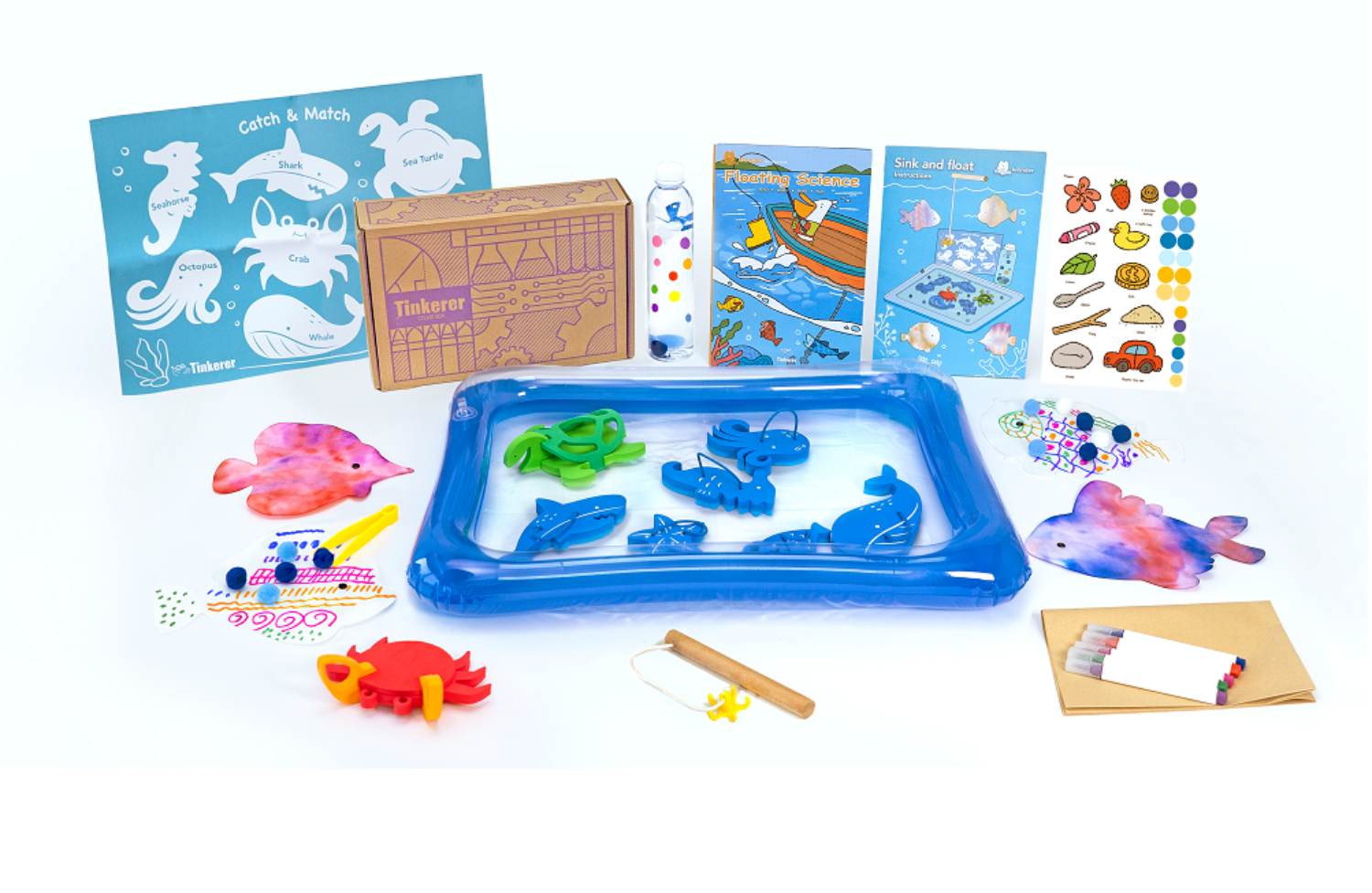 A children's activity kit with an ocean theme is displayed on a white background. The kit includes a blue inflatable water table containing several wooden sea creature shapes, a small wooden fishing rod, illustrated activity sheets featuring sea animals and experiments about floating and sinking, a box labeled Tinkerer, a bottle with colored liquids, a sheet of stickers with various small images, a set of markers, and several pieces of decorated paper or cardstock crafted to resemble fish and a crab. The arrangement suggests a comprehensive set designed for educational play and creative activities related to marine life.