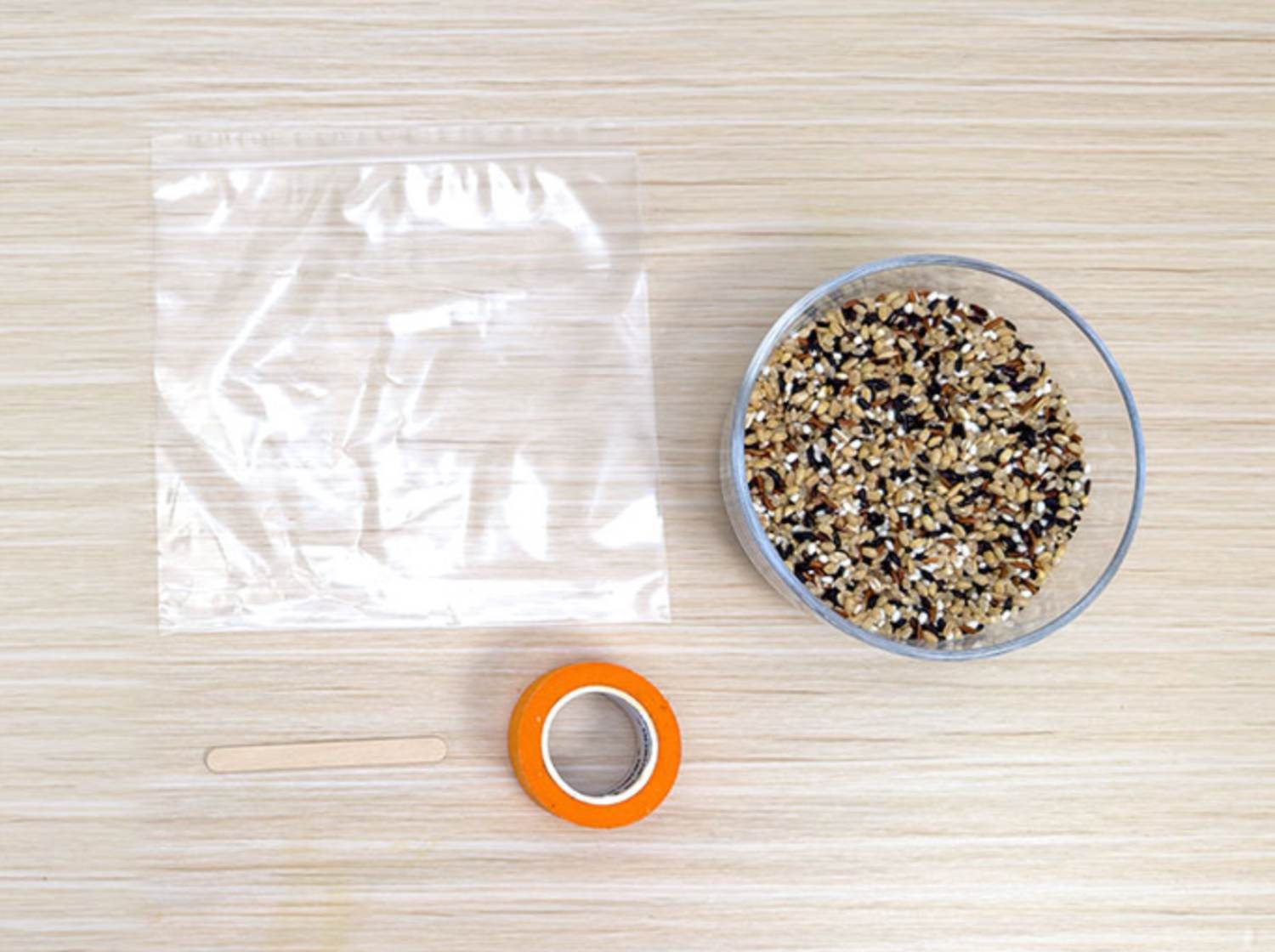 Arranged on a light wood-grain surface are a few simple items, suggesting preparation for a craft or sensory activity. A clear plastic zip-top bag lies flat, beside a small, clear bowl containing mixed grains of rice. A roll of orange masking tape and a small wooden craft stick are also present. The scene implies a DIY project, possibly involving filling the bag with rice to create a sensory toy or weighted object.