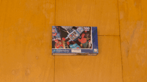 A jigsaw puzzle box sits on a golden-yellow surface, likely a wooden floor or table. The box features a picture of colorful vintage toys, including a prominent blue robot. The box indicates it contains 500 pieces. The camera focuses on the box, which remains stationary throughout the video.