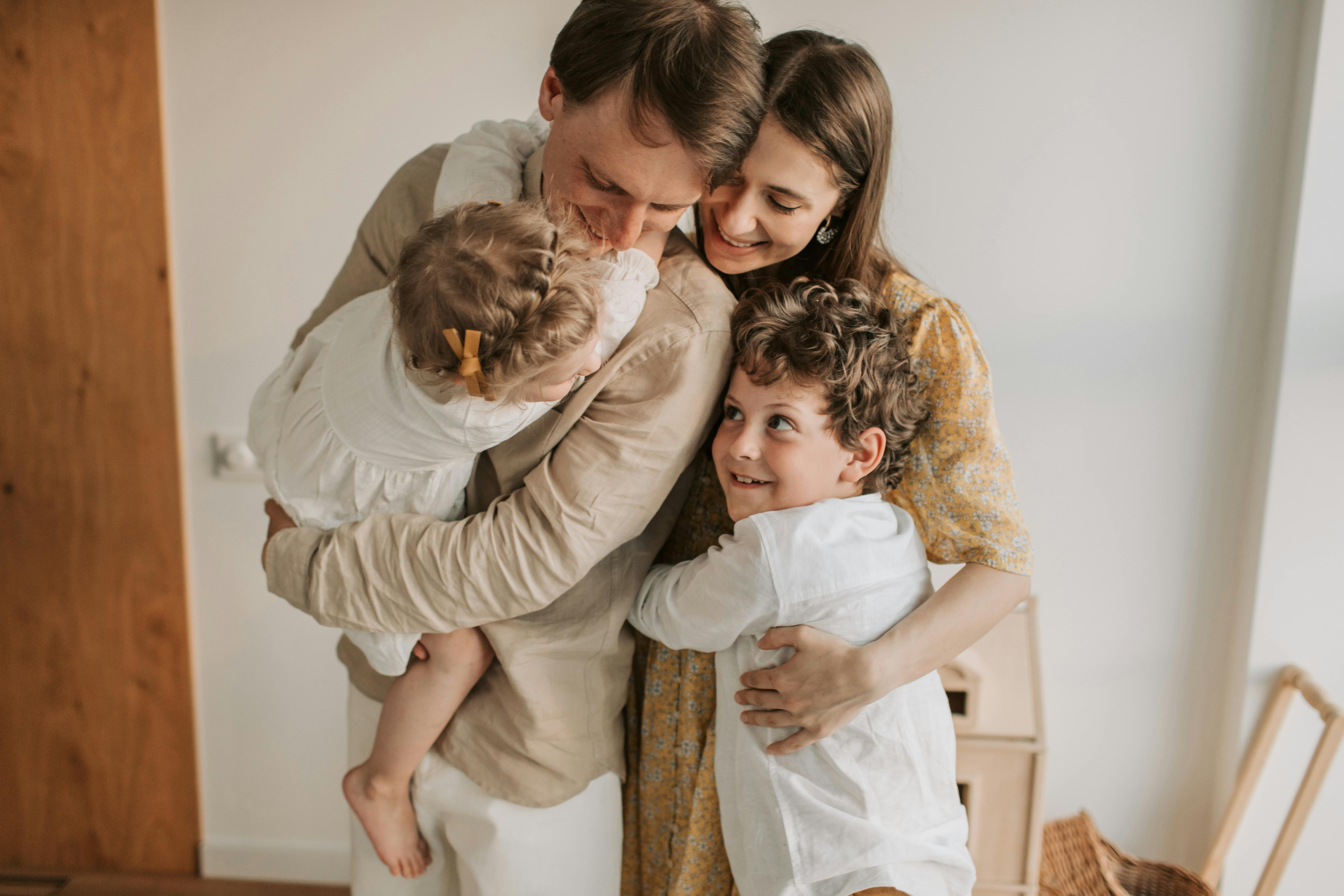A happy family of four shares a warm and affectionate group hug in a brightly lit room. The father, wearing a beige shirt, holds their toddler daughter in a white dress and lovingly nuzzles her. The smiling mother, in a yellow floral dress, embraces her husband and their young son. The curly-haired son, wearing a white shirt, looks up with a joyful expression while hugging his parents.