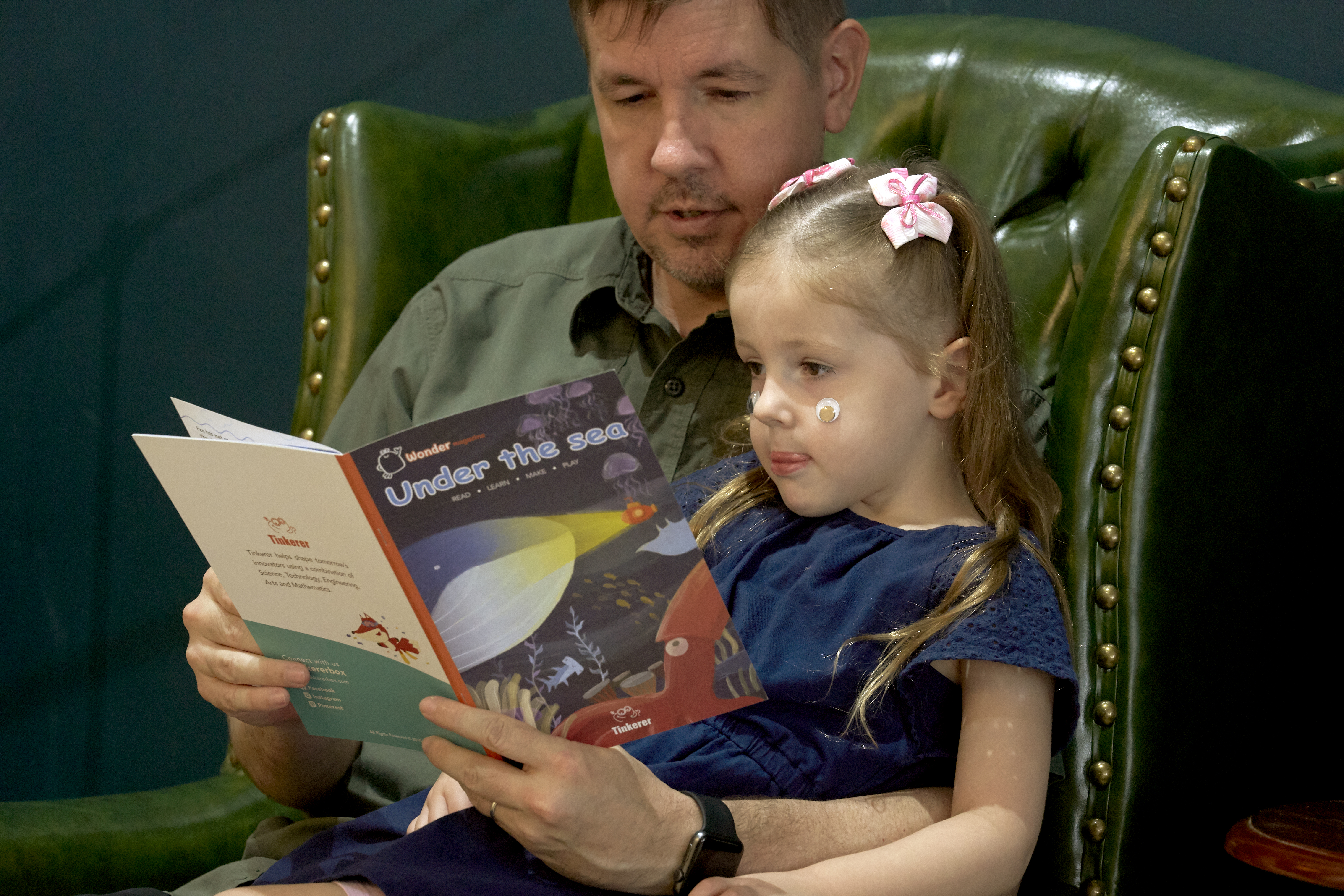 A father sits in a green leather armchair, reading a book titled Under the sea to his young daughter, who is nestled on his lap. The girl, wearing a blue dress and pink hair bows, listens attentively with a sticker on her cheek.A father sits in a green leather armchair, reading a book titled 
