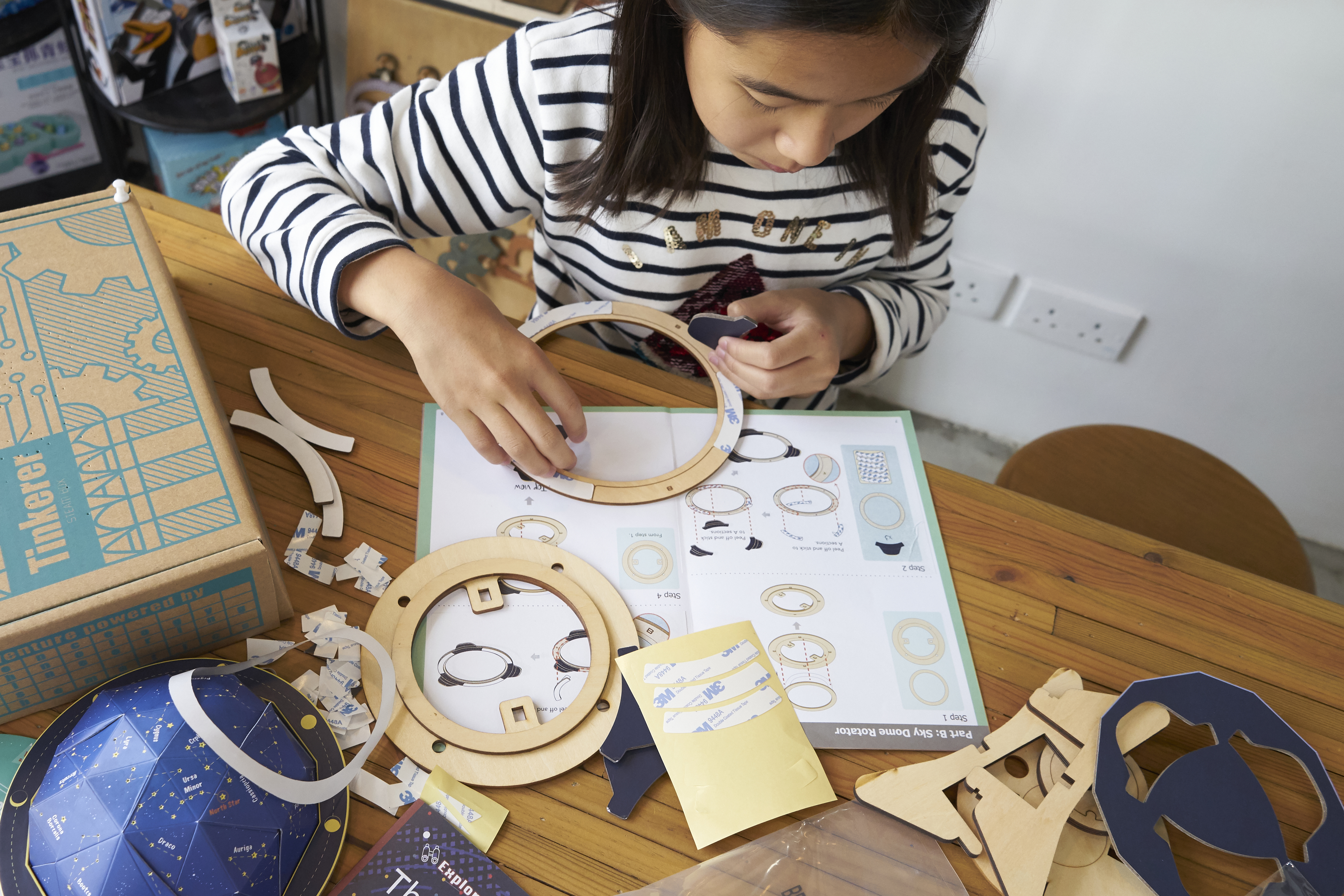 A young girl wearing a striped shirt sits at a wooden table, concentrating on assembling a science kit. The table is covered with wooden rings, paper cutouts, adhesive strips, and an instruction booklet. A partially completed blue dome with star constellations and a Tinkerer brand box are visible, indicating an astronomy-themed project.