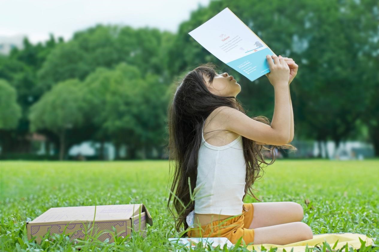 In a sun-dappled meadow, a young girl finds a moment of pure bliss. Her long dark hair cascades down, contrasting with her crisp white tank top and cheerful yellow shorts. With playful ingenuity, she holds a book aloft, creating an impromptu sunshade. Beneath this literary umbrella, her face radiates unbridled joy, her smile as bright as the day itself. The setting is a verdant paradise - lush grass carpets the ground, while trees stand sentinel in the background, their leaves whispering in the summer breeze. This snapshot captures the essence of carefree childhood summers, where simple pleasures and nature's beauty intertwine to create magic.