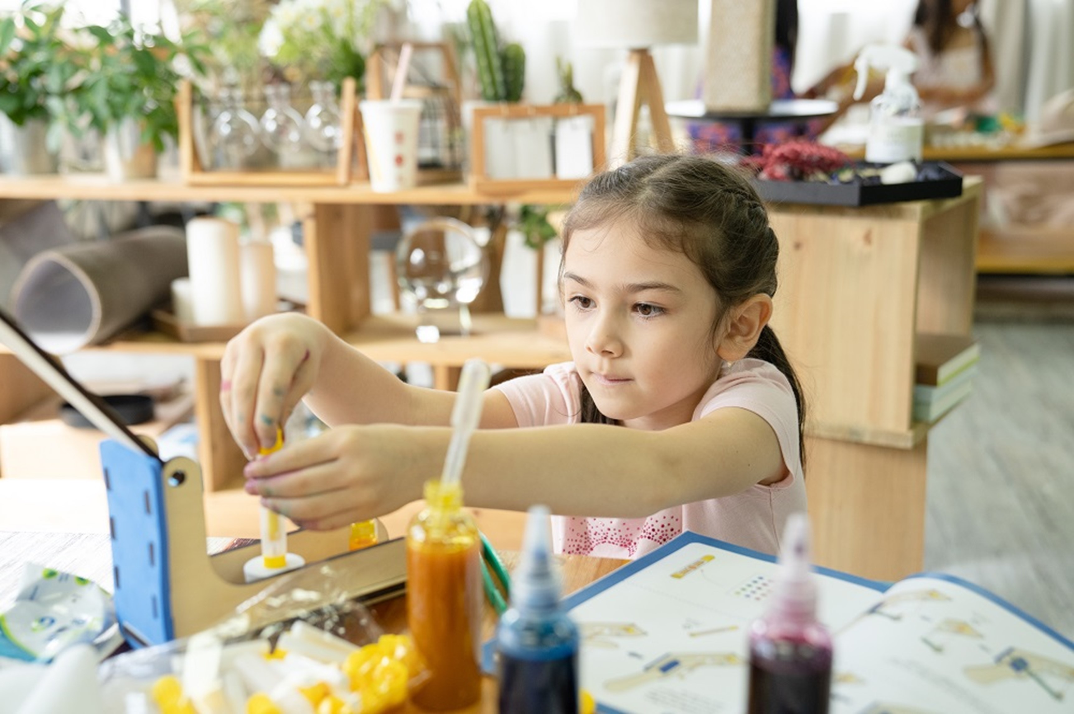 The image depicts a young girl engaged in a hands-on, creative activity within a vibrant, art-filled STEAM environment. The girl appears focused and intent as she works with various materials, including paint bottles and a drawing pad, suggesting an artistic or educational project.