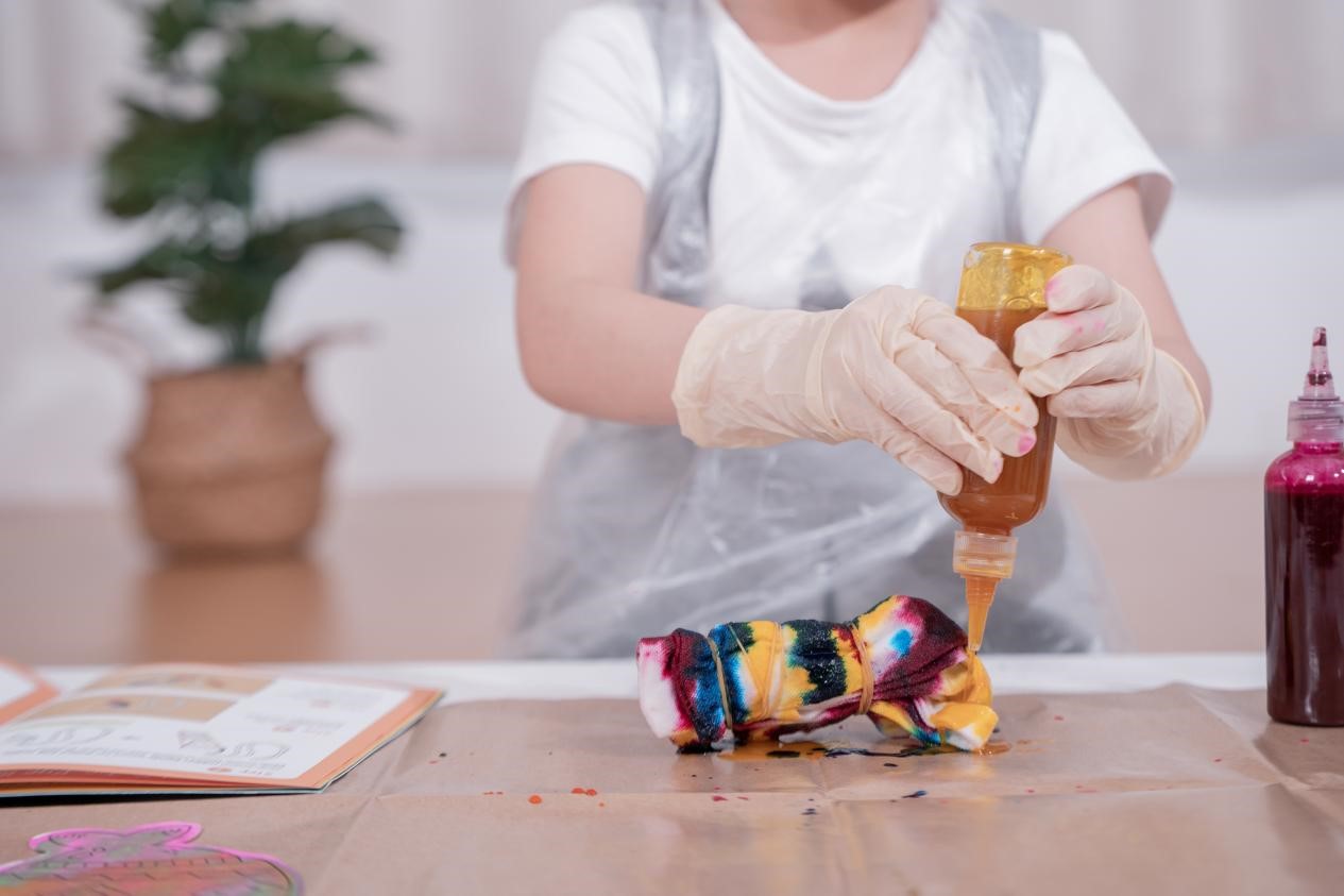 The image depicts a person's hands engaged in an artistic, hands-on activity, using various materials such as a colorful fabric and a bottle of liquid. The person appears to be in a creative STEAM workspace, surrounded by plants and other artistic elements, suggesting an environment conducive to exploration and self-expression. The focused attention and the array of materials indicate this is likely an artistic and educational endeavor, where the individual is immersed in a process of experimentation, discovery, and the blending of creative and technical skills.