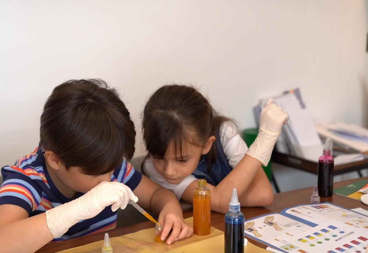 The image depicts two young children intently focused on a hands-on learning activity, their faces bent over a table filled with various art supplies and materials. The children's expressions convey a sense of concentration and engagement as they work together, suggesting an environment conducive to collaborative exploration and discovery. The setting, with its array of colorful bottles and drawing implements, evokes a creative, educational STEAM space where children are encouraged to experiment, problem-solve, and tap into their innate curiosity and imagination. This scene captures a moment of active, immersive learning, where the children appear to be fully immersed in the process of creating, investigating, and expanding their understanding of the world around them.