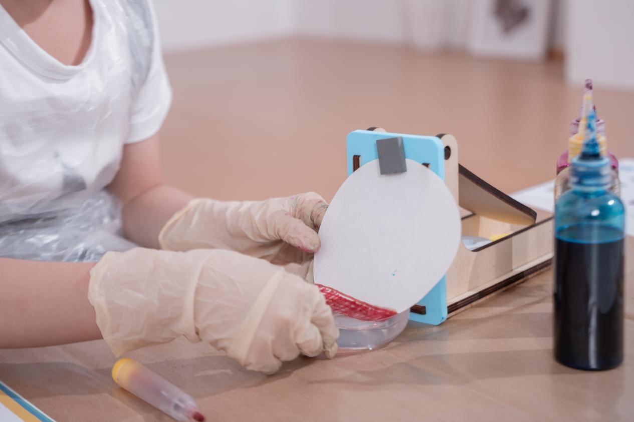 A person's hands engaged in a hands-on, creative activity using various tools and materials. The person is wearing protective gloves, suggesting a focus on safety and precision as they manipulate a device that appears to be a cutting or printing tool. The setting includes additional art supplies, such as colored bottles, indicating an environment conducive to artistic exploration and experimentation. The person's posture and the tools at hand suggest they are immersed in a process of creating, problem-solving, or learning through a hands-on, tactile approach. This scene captures a moment of focused, engaged learning or creative expression, where the individual is actively participating in a process that blends technical skills with artistic expression.