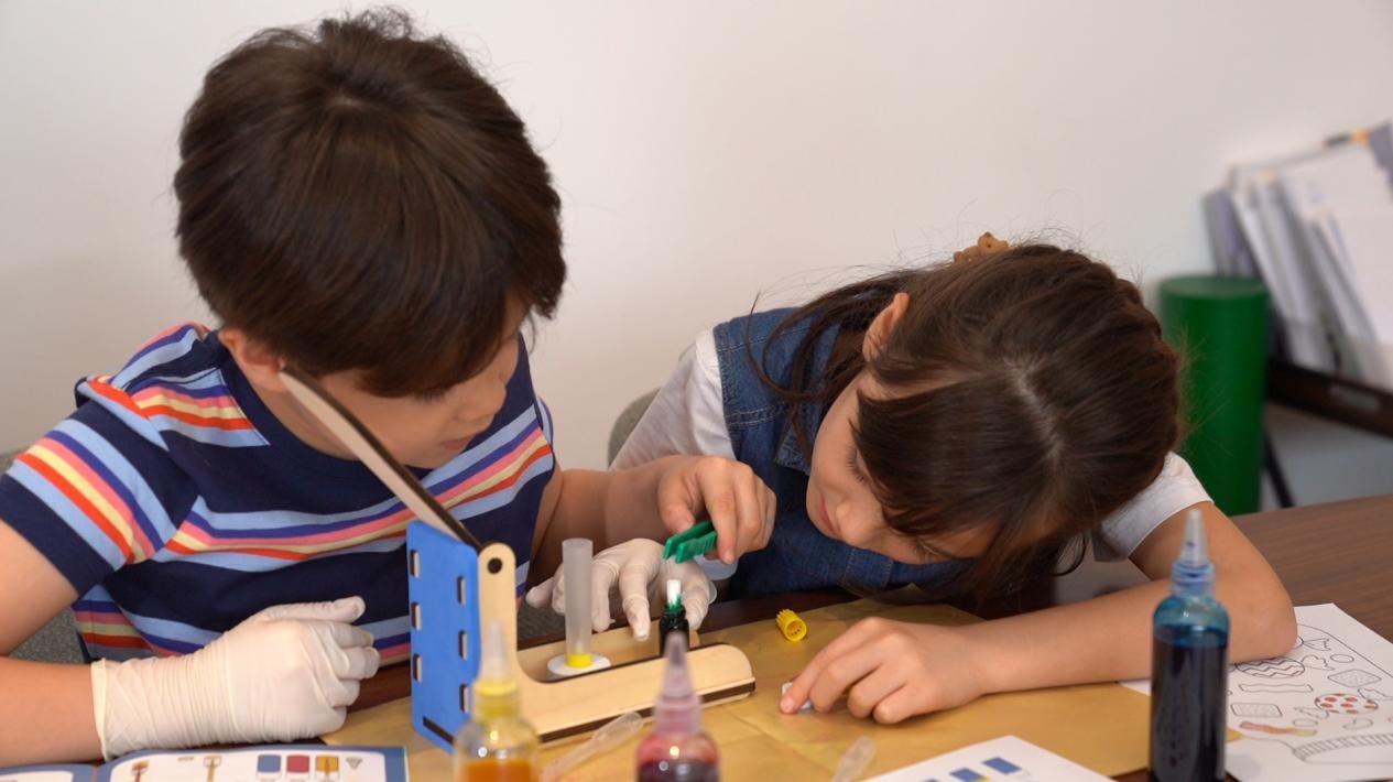 The image depicts two young children intently focused on a hands-on, educational STEAM activity, their faces focused over a table filled with various art supplies and materials. The children's expressions convey a sense of deep concentration and engagement as they work together, suggesting an environment conducive to collaborative exploration and discovery. The setting, with its array of colorful tools and implements, evokes a creative, learning-oriented space where children are encouraged to experiment, problem-solve, and tap into their innate curiosity and imagination. This scene captures a moment of active, immersive learning, where the children appear to be fully immersed in the process of creating, investigating, and expanding their understanding of the world around them.