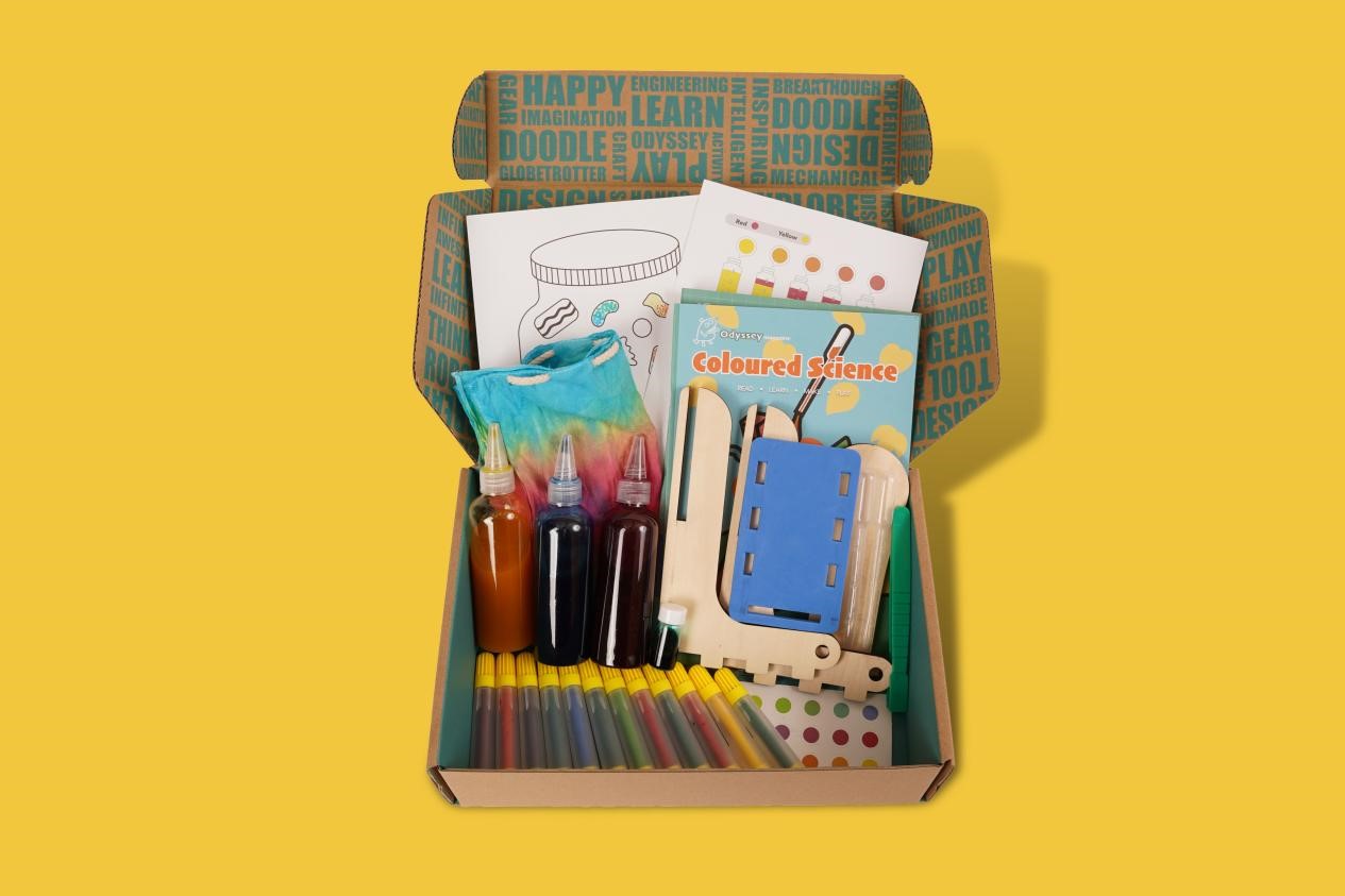 This image showcases a vibrant, engaging STEM-themed activity box or educational kit. The box is filled with an array of colorful, hands-on materials and tools designed to spark creativity, imagination, and scientific exploration. The contents include art supplies like markers, paints, drawing materials and a book titled Coloured Science. This diverse collection of items suggests a multidisciplinary approach, blending artistic expression with scientific concepts and problem-solving.