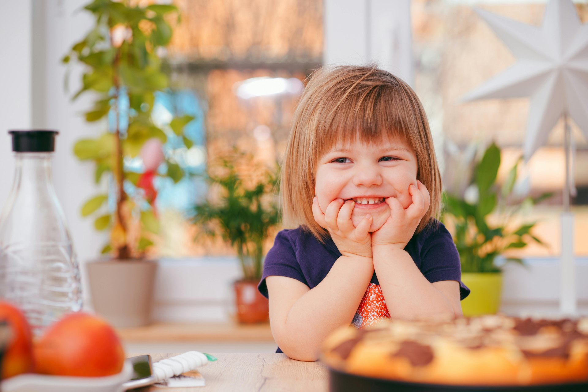 A happy young girl with short, light-brown hair and bangs, wearing a purple t-shirt, rests her chin on her hands and smiles while sitting at a wooden table. In front of her is a blurry marble cake, and in the background are potted plants and a white star decoration near a window.