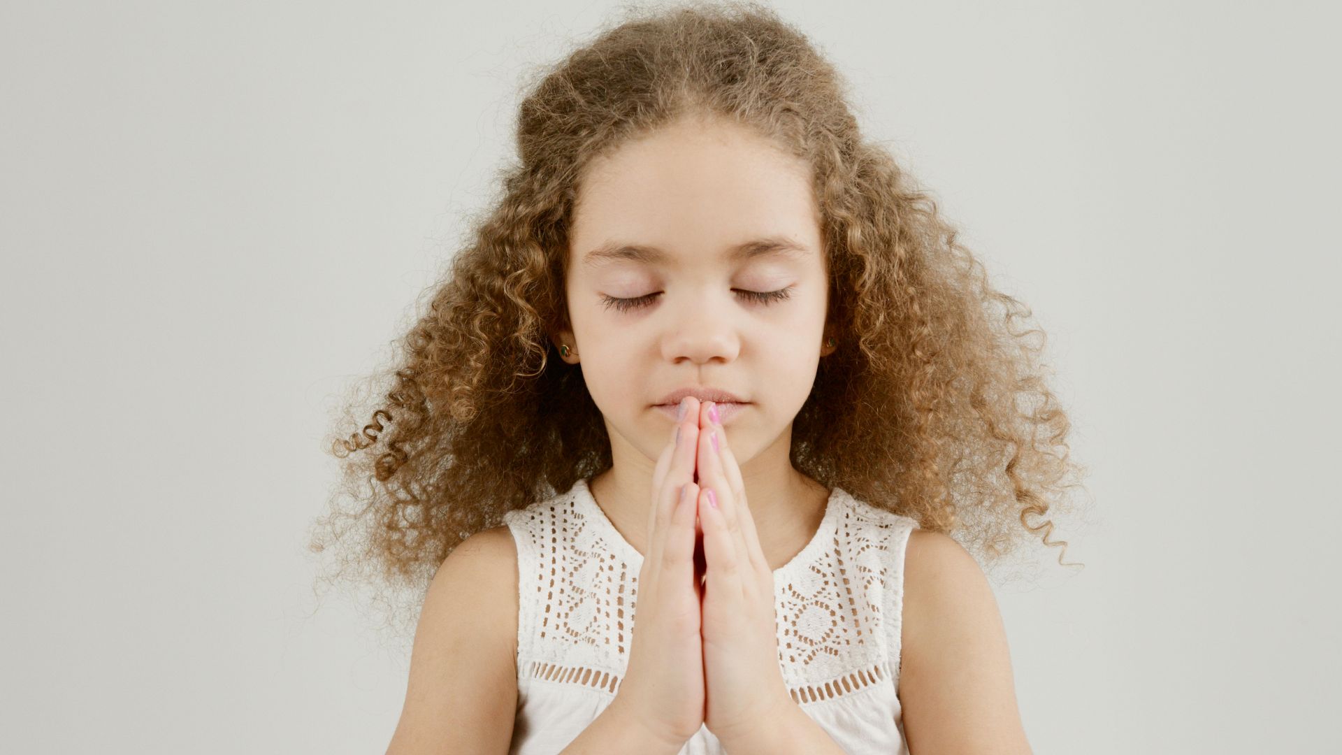 A young girl with long, curly, light-brown hair has her eyes closed and hands pressed together in prayer. She is wearing a white lace top and is positioned against a plain, light-gray background.