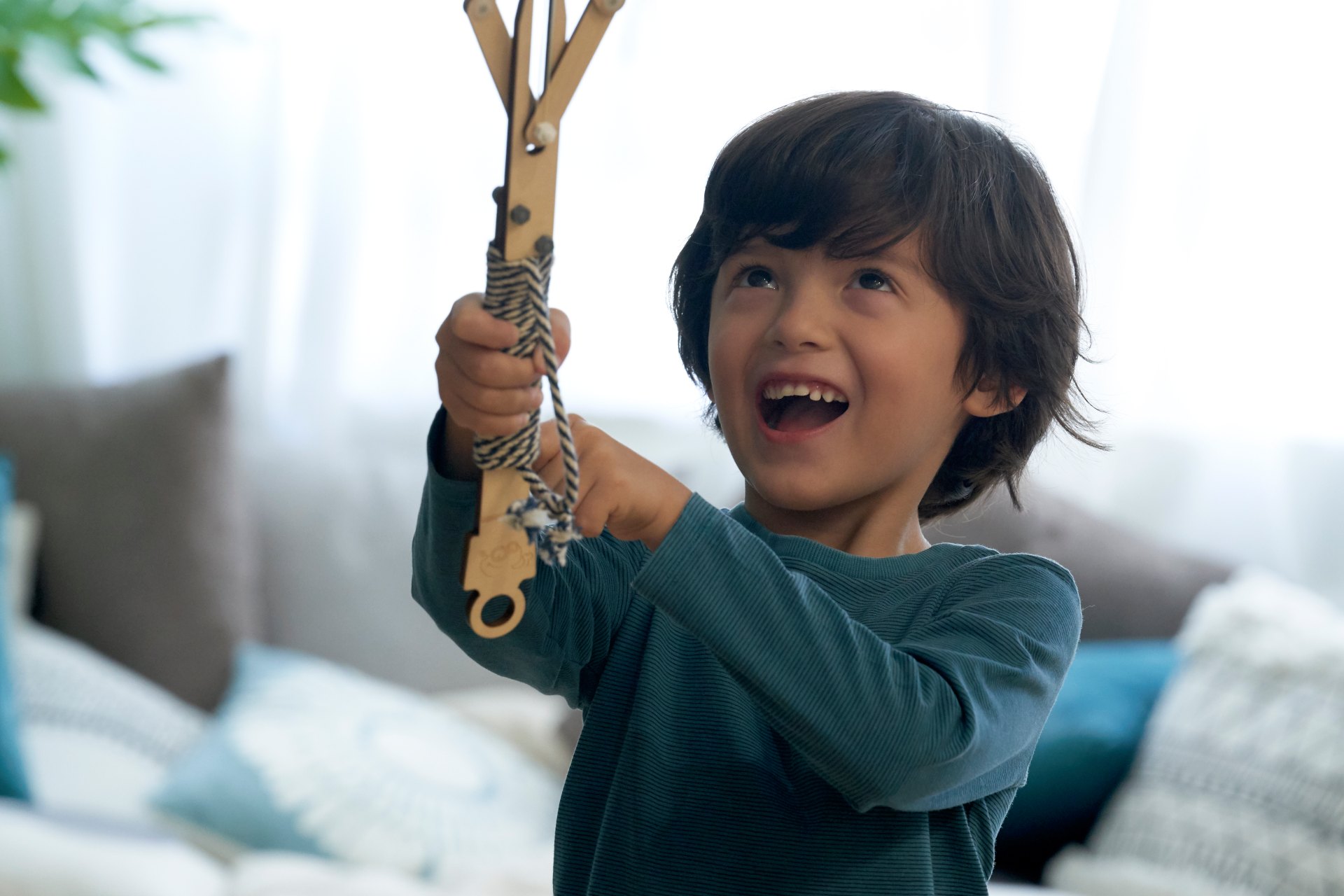 A young boy with dark wavy hair and a joyful expression looks up with his mouth open in excitement as he holds up a wooden toy. He is wearing a long-sleeved, dark teal striped shirt, and the background is a brightly lit, out-of-focus living room.